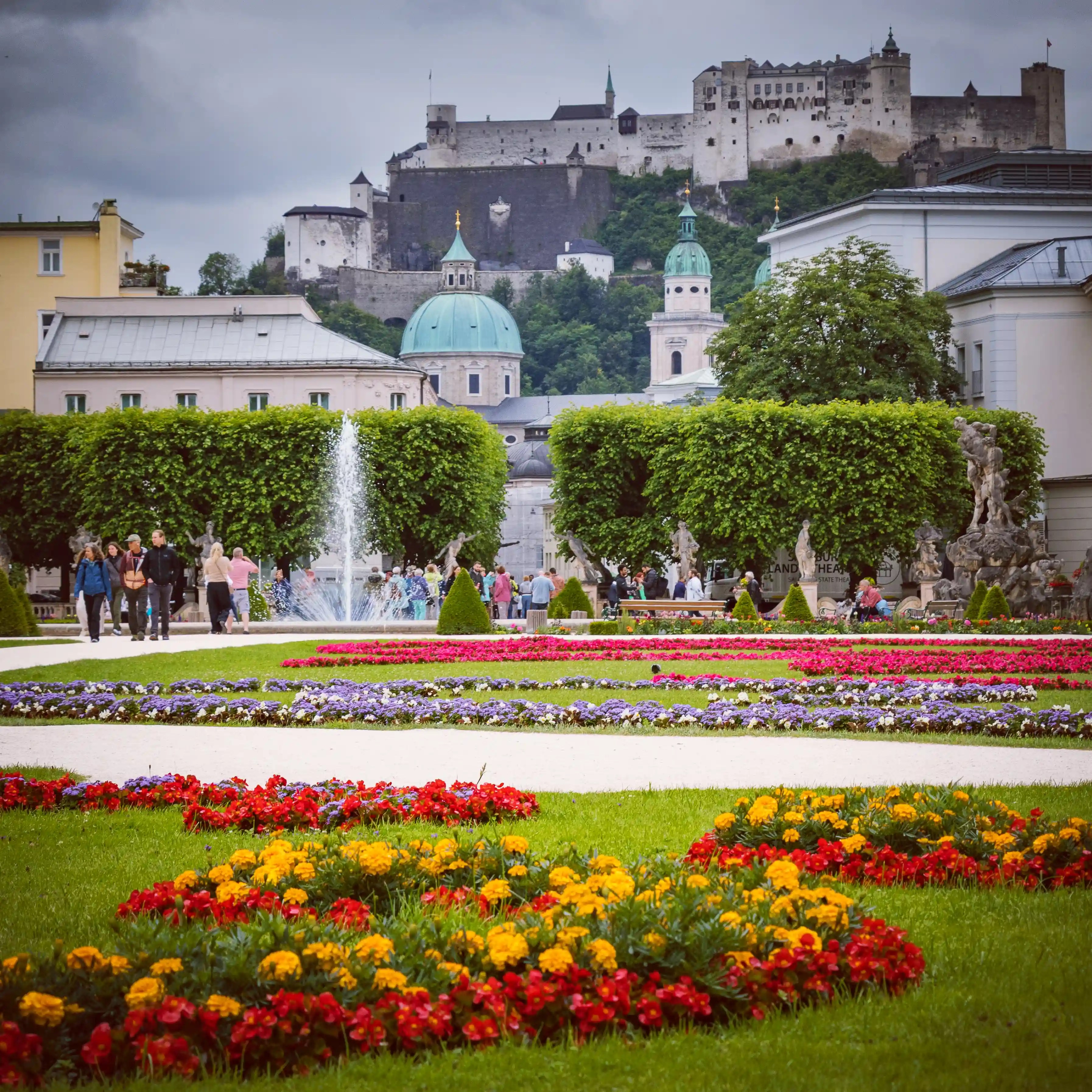 Bright flowerbeds surround a central fountain as visitors walk through Mirabell Gardens, with Hohensalzburg Fortress visible in the background.