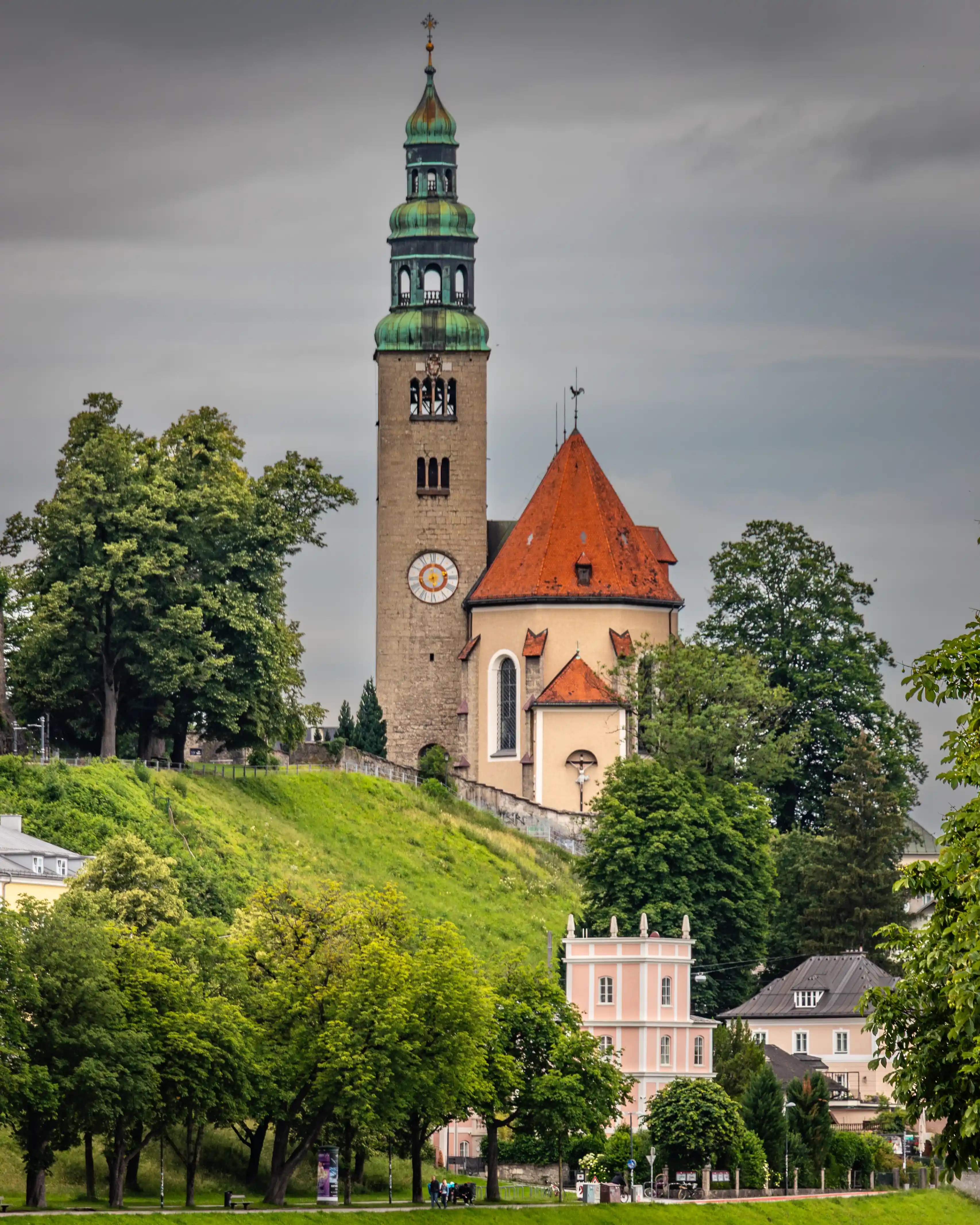 A tall stone church tower with a green copper spire and clock rises above a grassy hillside, with trees and pastel buildings at the base in Salzburg.