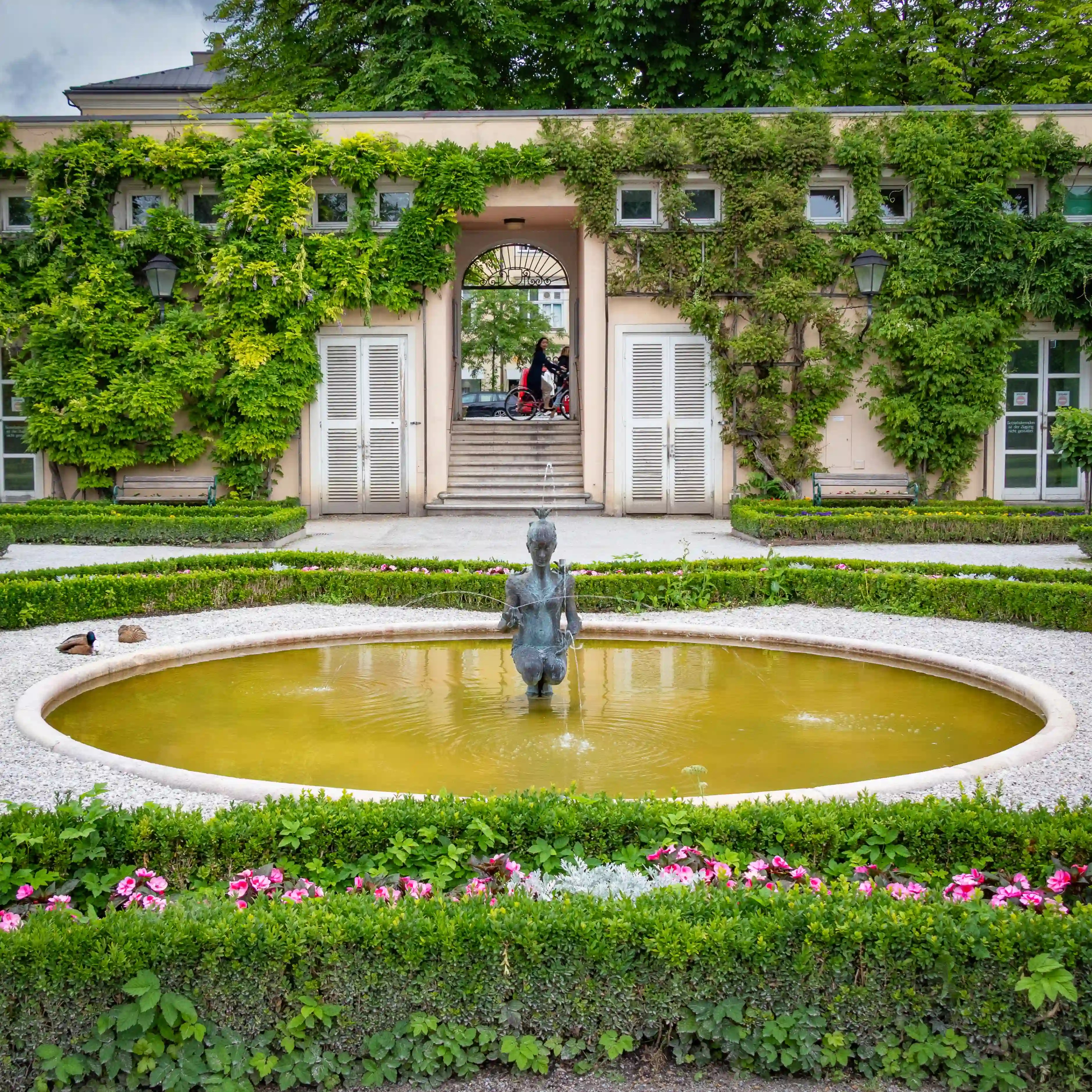 A small bronze fountain statue stands in a circular pool, framed by trimmed hedges and an ivy-covered wall at Mirabell Gardens.
