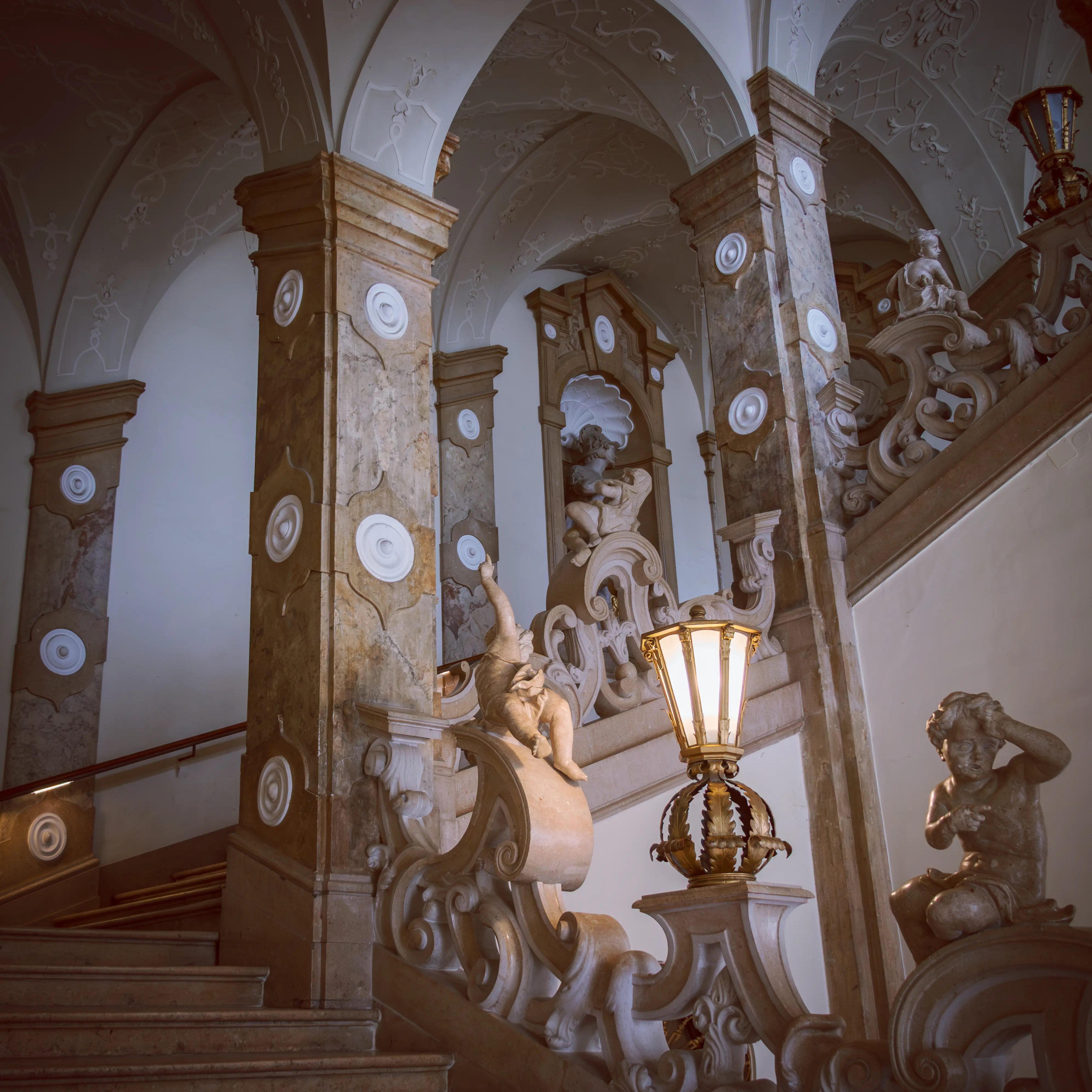 An ornate marble staircase inside Mirabell Palace, decorated with sculpted cherubs, stone columns, and a glowing lantern.