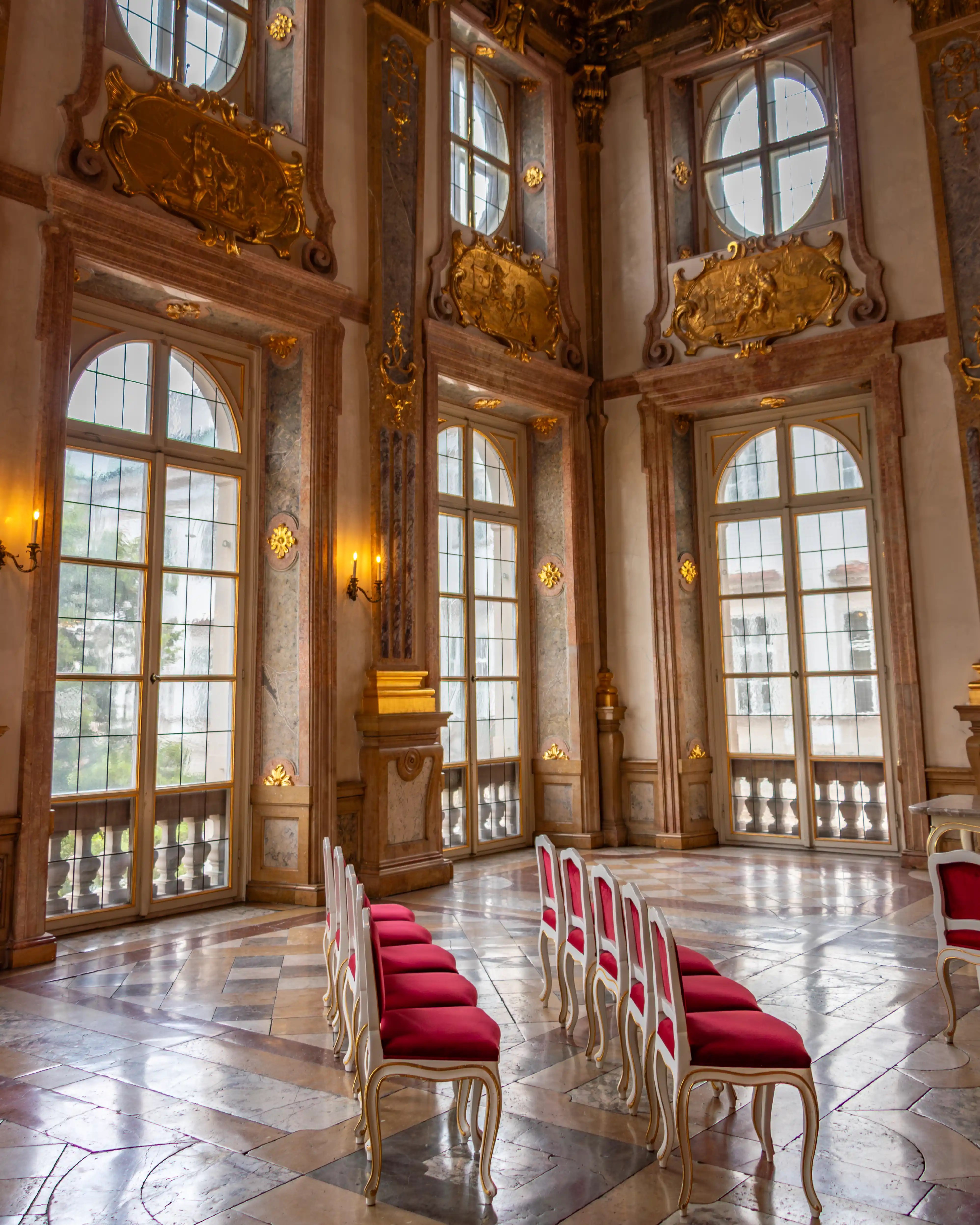 A grand hall with tall arched windows, gilded wall panels, marble floors, and rows of red chairs arranged inside Mirabell Palace.