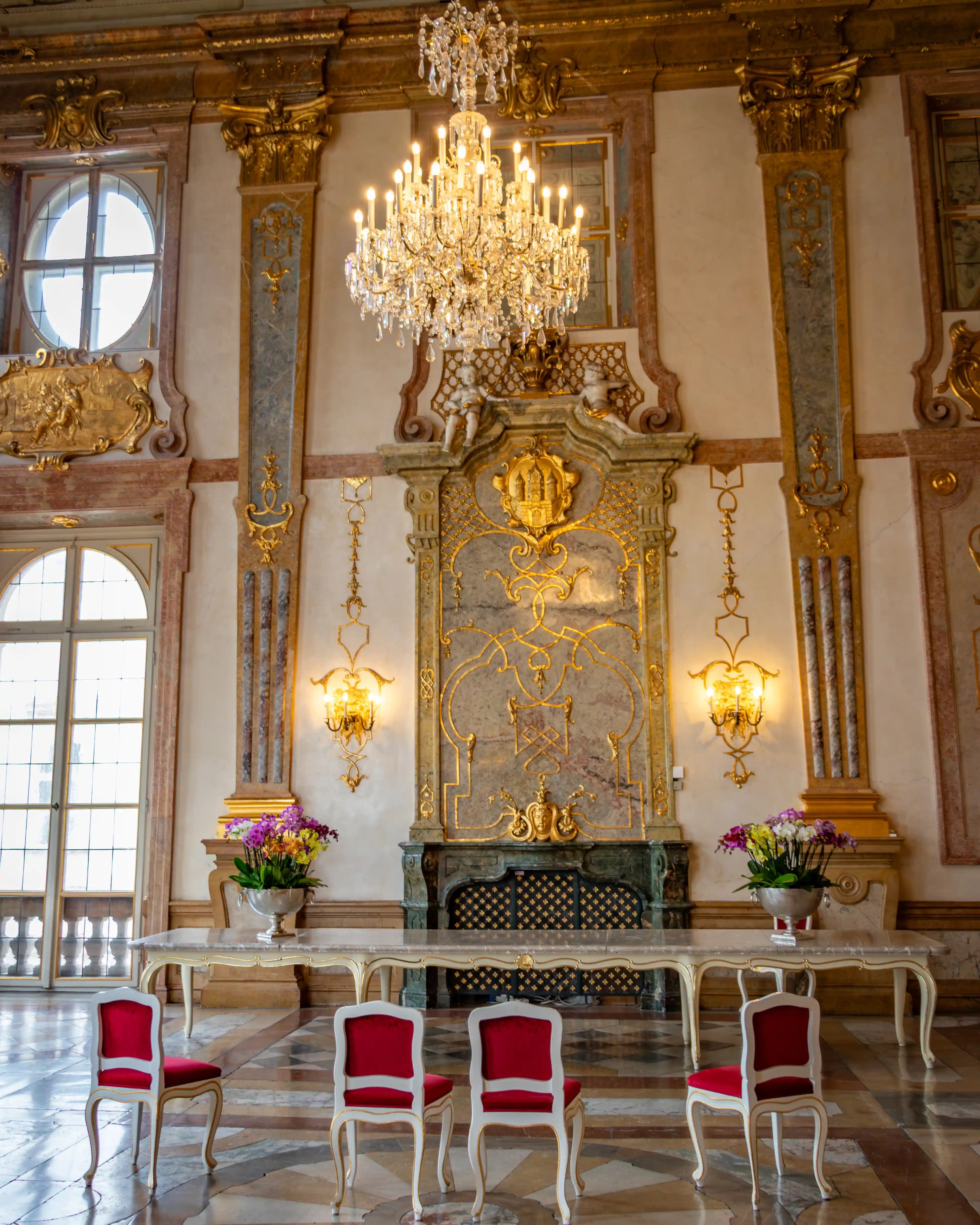 A richly decorated wall inside Mirabell Palace with gold detailing, a crystal chandelier, tall windows, and red upholstered chairs below.