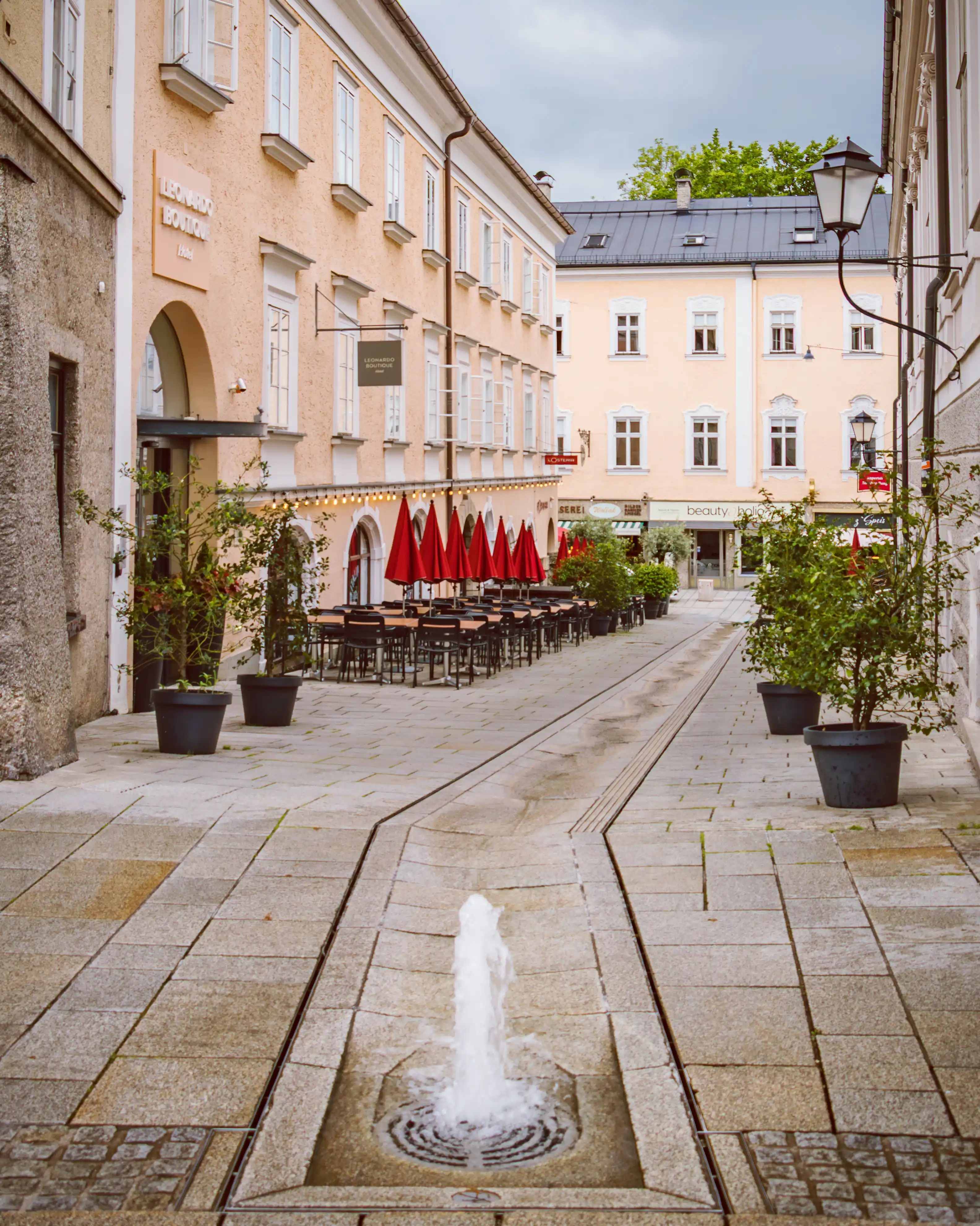 A narrow paved lane with a small ground-level fountain leading toward pastel-colored buildings with café tables and red umbrellas in Salzburg.