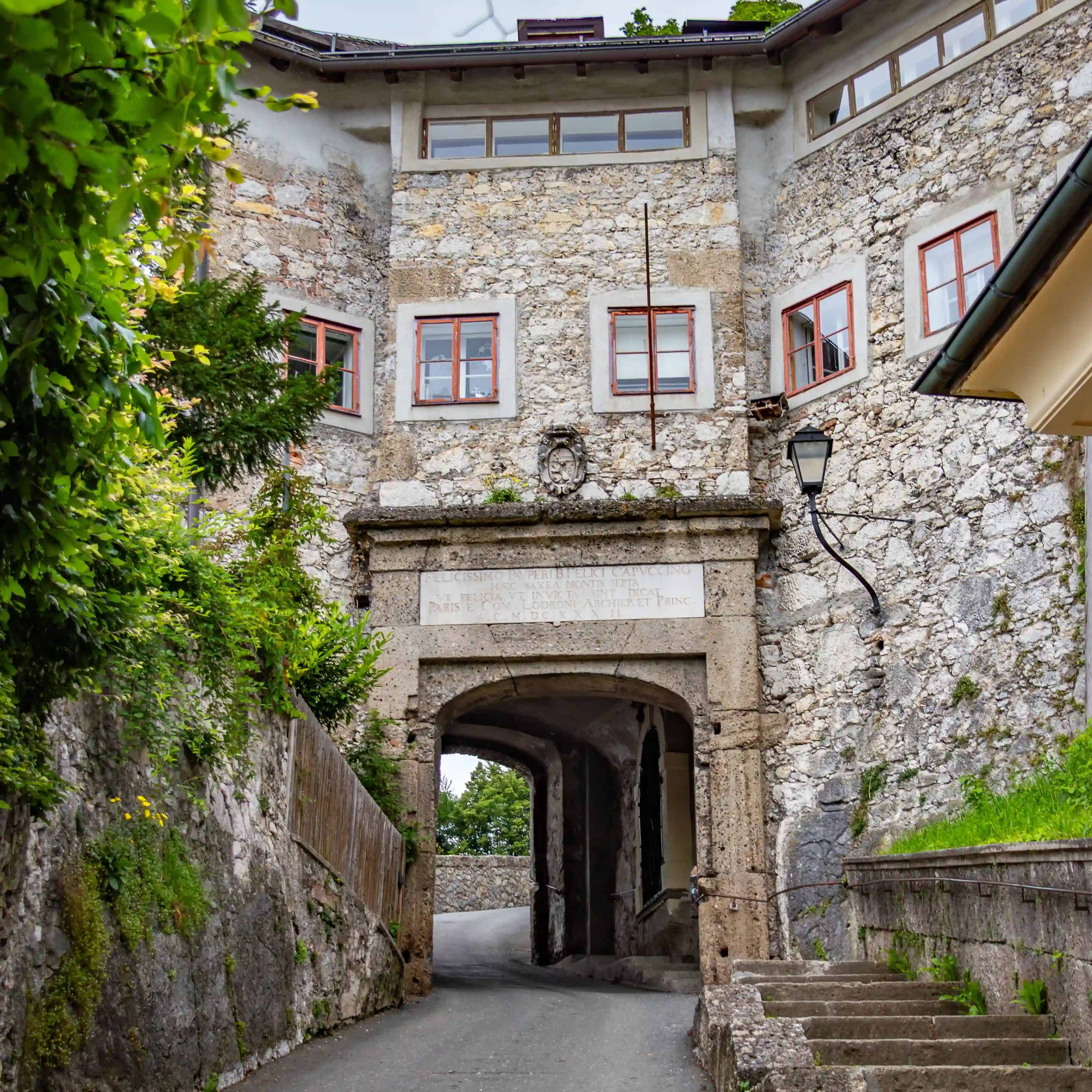 A narrow stone archway built into a rough limestone wall opens onto a paved path on Kapuzinerberg in Salzburg.