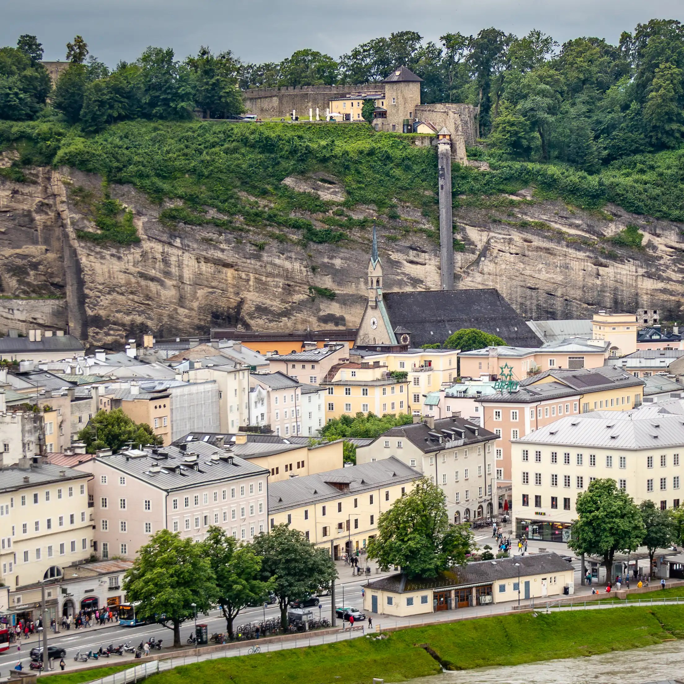A view over Salzburg’s Old Town showing pastel historic buildings along the Salzach River with St. Blasius Church and the steep rock face of the Mönchsberg rising behind it.