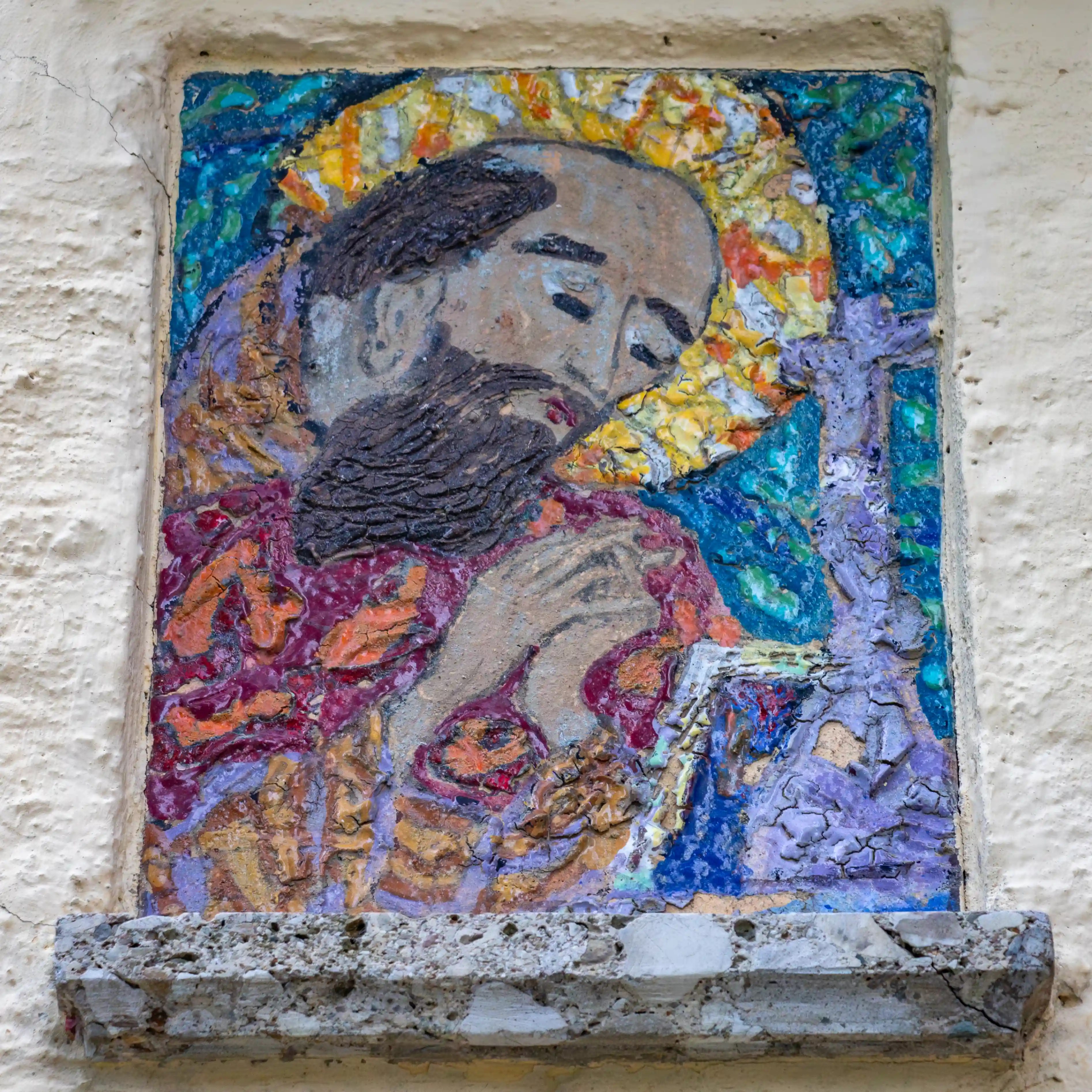 A weathered religious painting set into a stone wall, showing a bearded figure with a halo in vivid blues and golds.