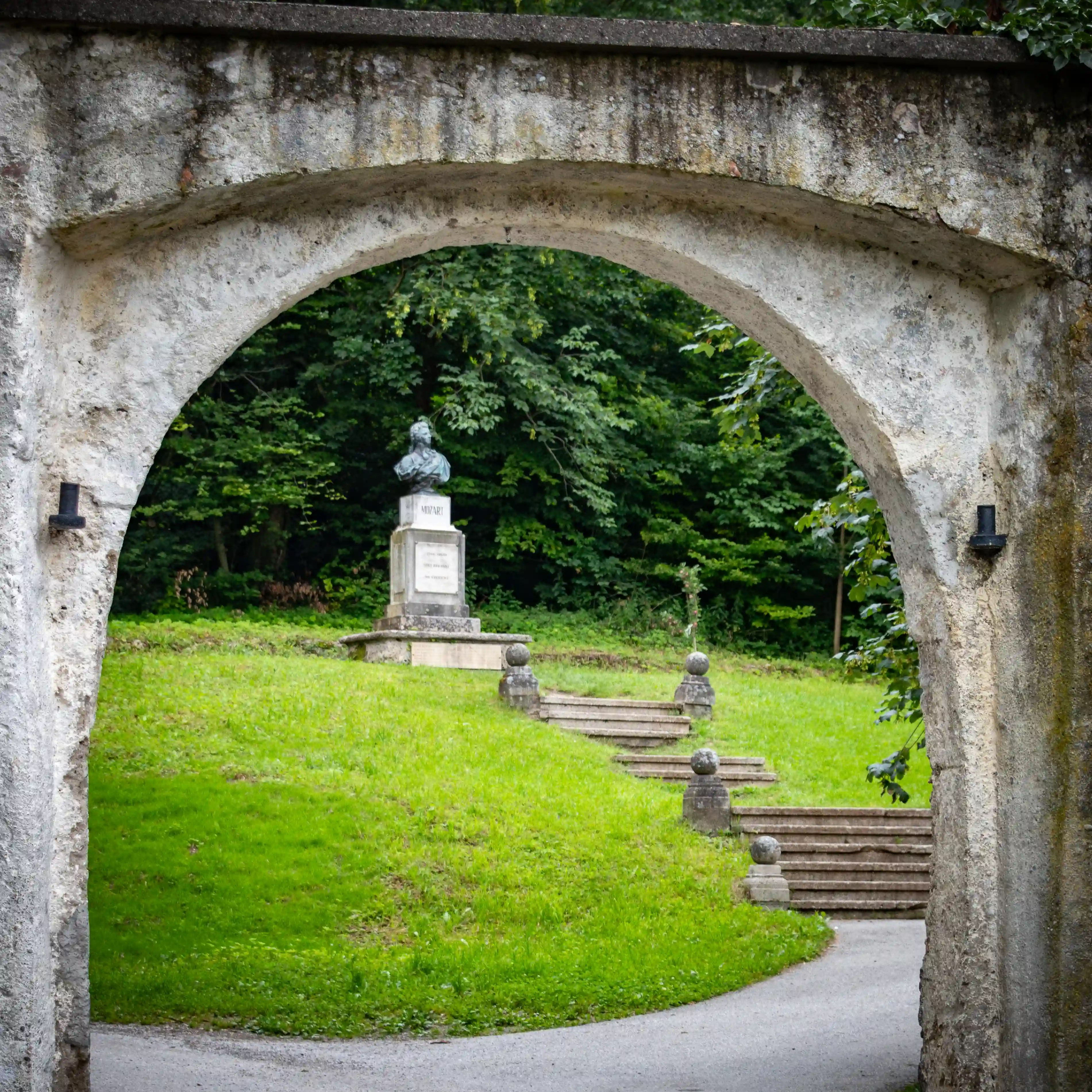 A stone archway framing a statue of Mozart set on a grassy hillside with steps leading up through the trees.