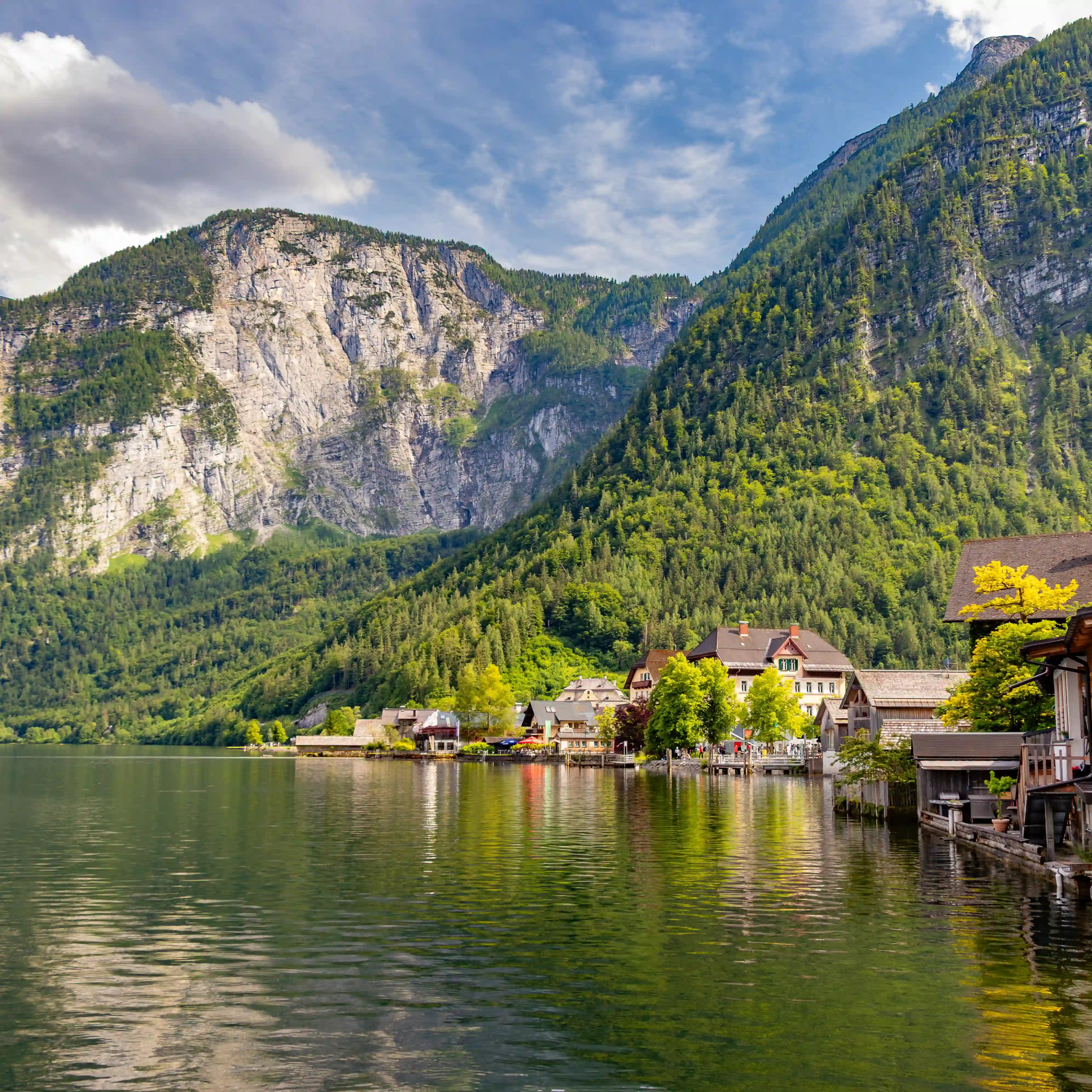 Lake Hallstatt with calm green water, village buildings along the shoreline, and steep rocky mountains rising behind them.