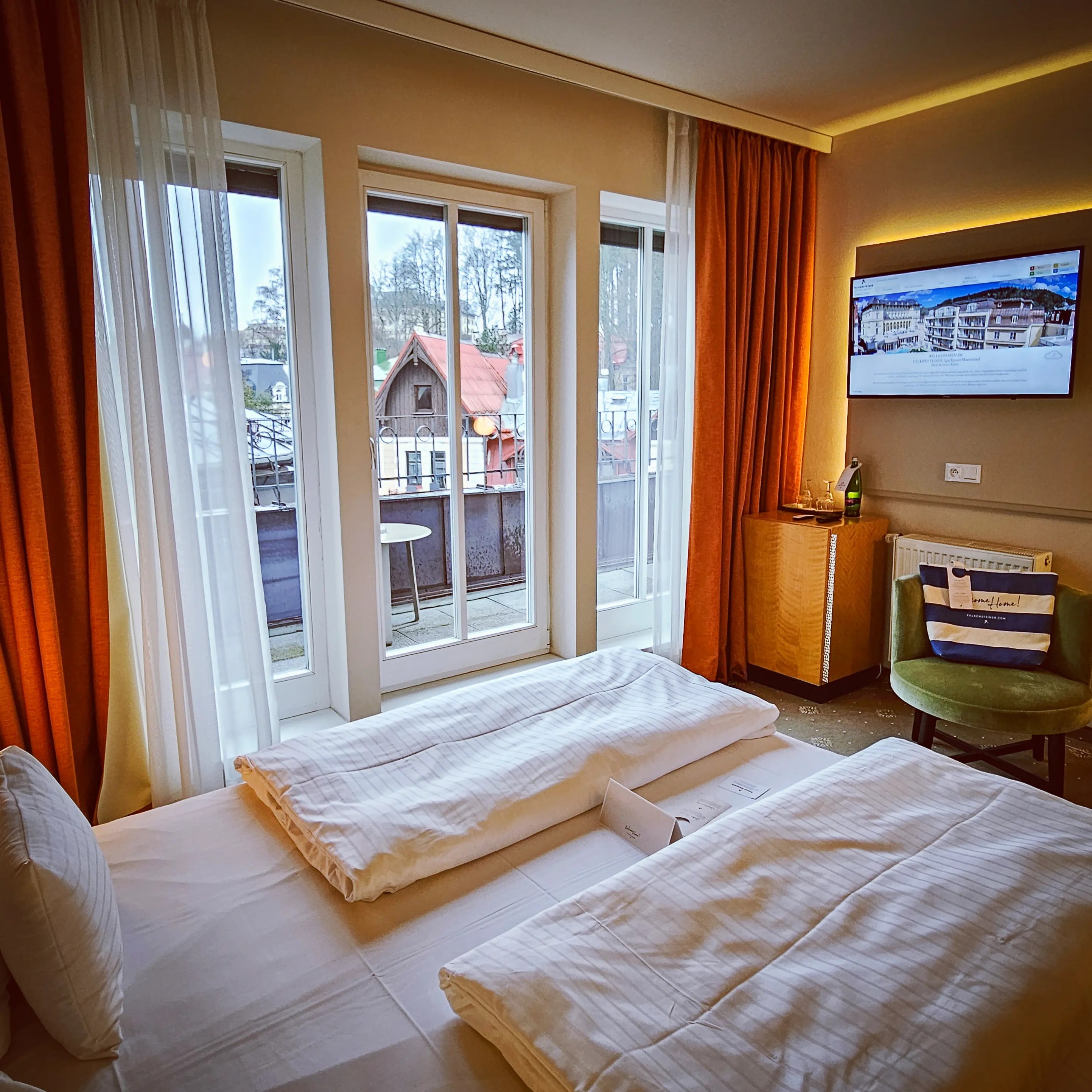 A modern hotel room with a neatly made bed, folded duvets, tall windows, and a balcony overlooking neighboring buildings in Mariánské Lázně.