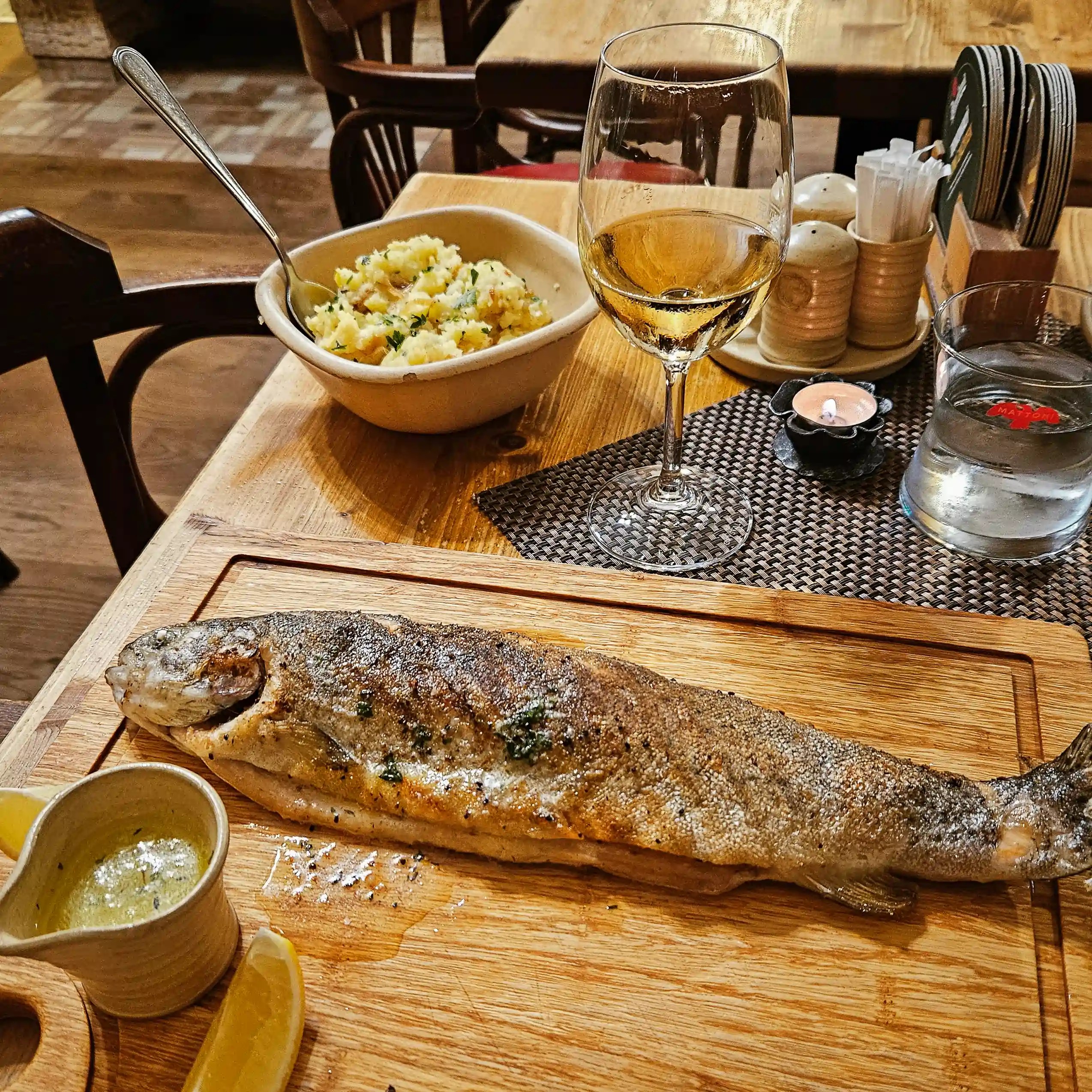 A whole grilled fish served on a wooden board with lemon wedges, herb butter, potatoes, and a glass of white wine.