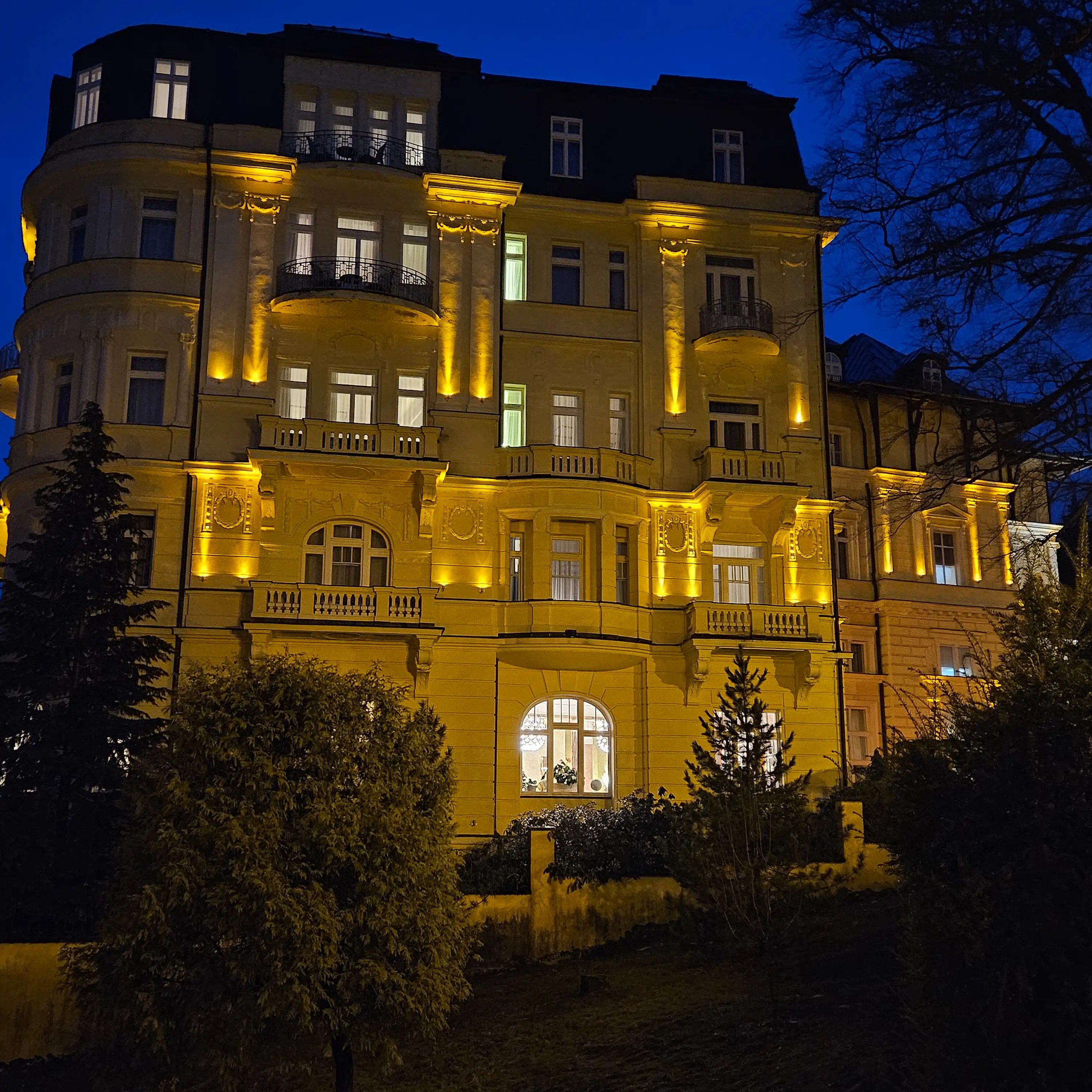 An elegant historic hotel building illuminated with warm golden lights against a dark blue evening sky in Mariánské Lázně.
