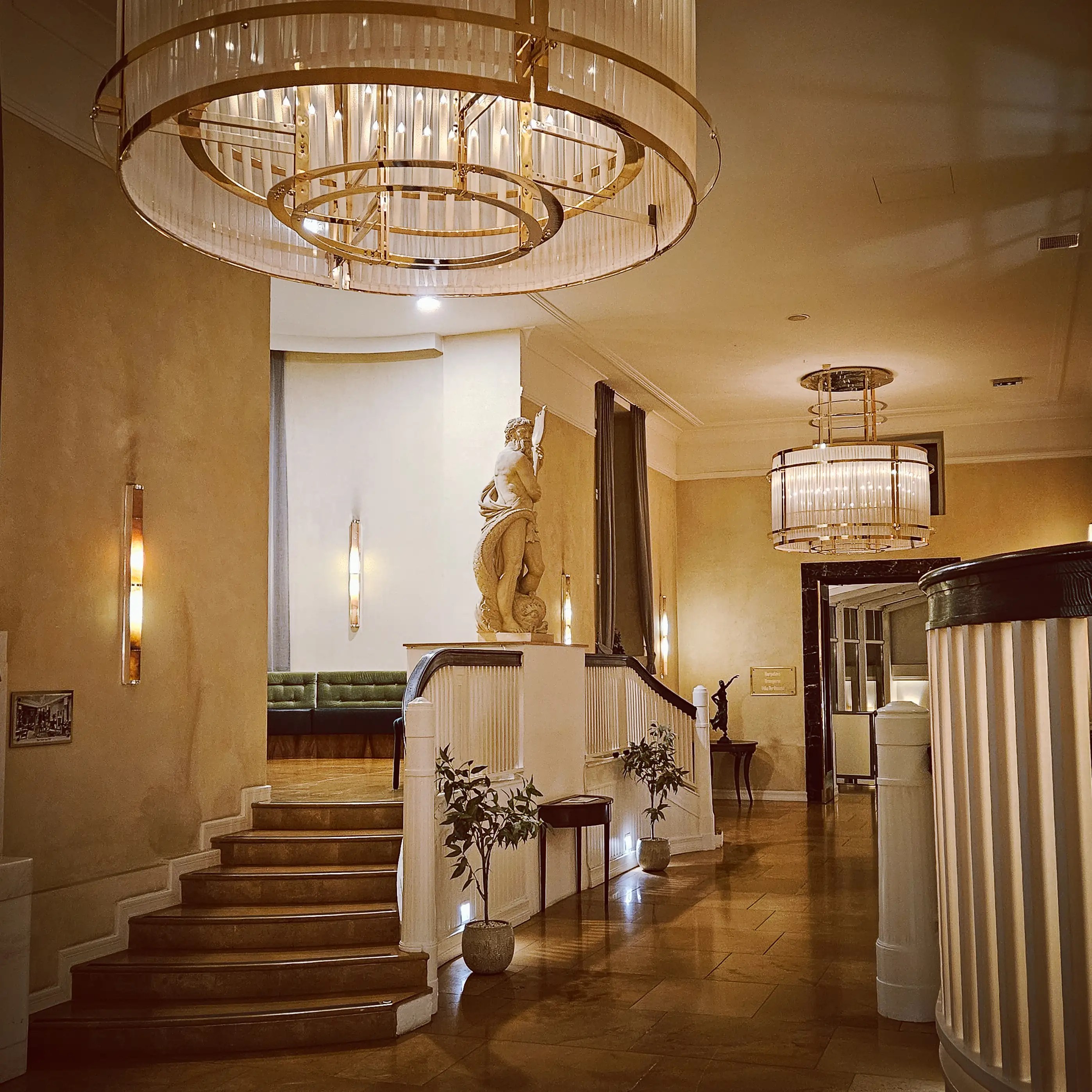 A grand interior staircase with curved railings, chandeliers, and a classical statue standing at the top landing inside Falkensteiner Spa Resort.