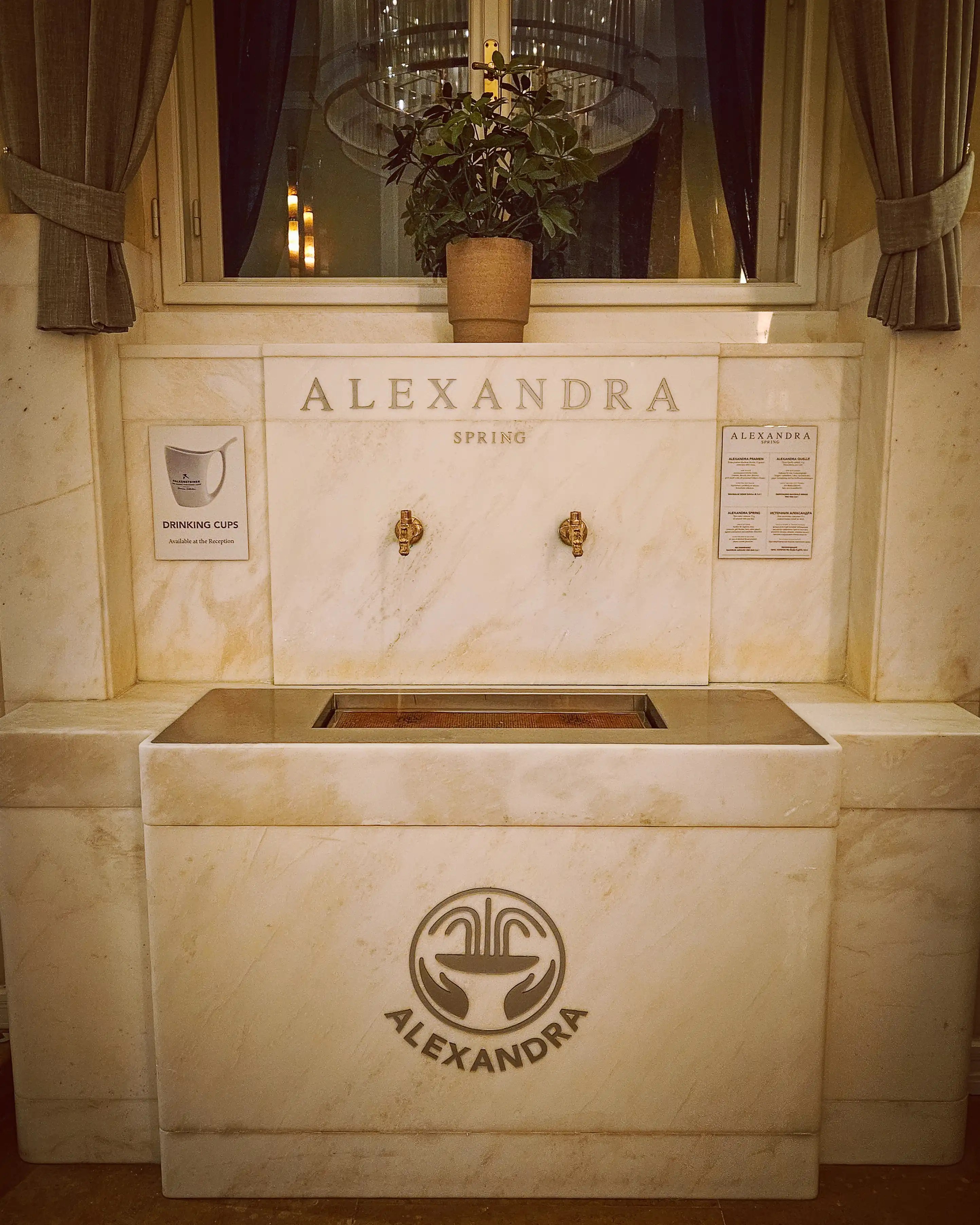 A marble drinking fountain labeled “Alexandra Spring” with two brass taps inside Falkensteiner Spa Resort.
