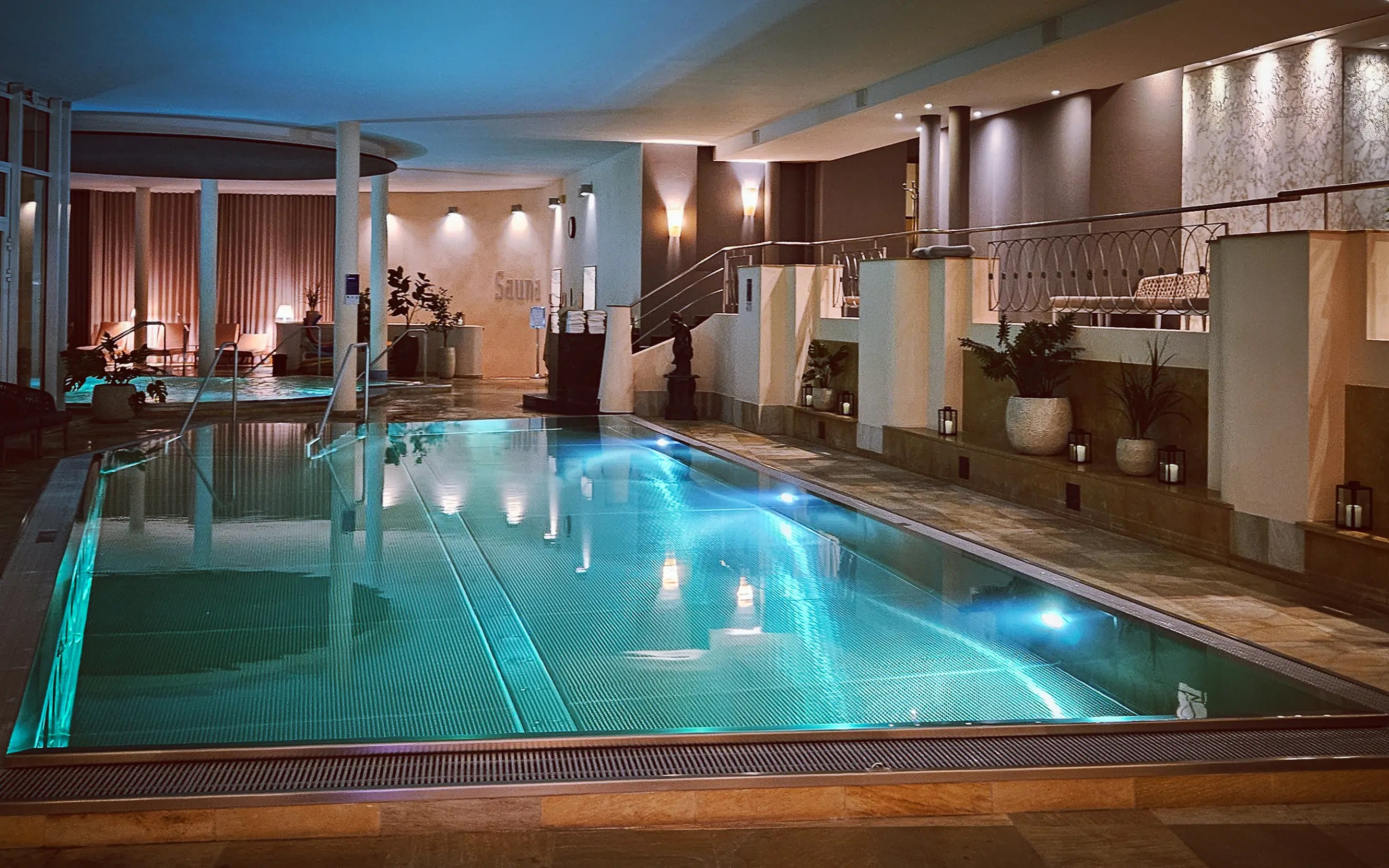 A softly lit indoor swimming pool with turquoise water, tiled floors, and lounge areas inside Falkensteiner Spa Resort.