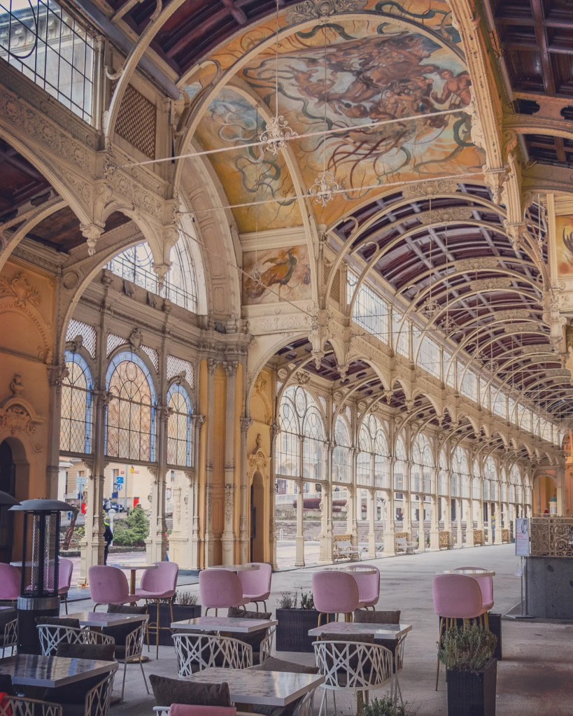 An ornate colonnade interior with arched windows, decorative columns, painted ceiling frescoes, and café tables with pink chairs in Mariánské Lázně.