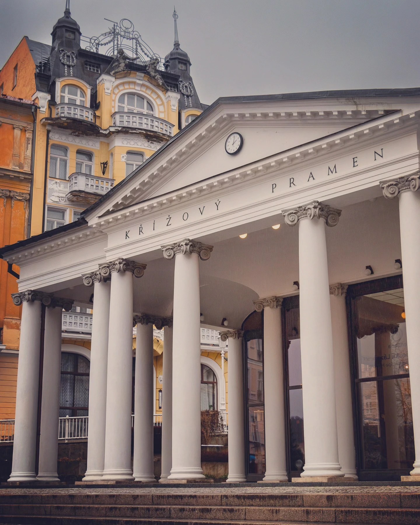 A neoclassical portico labeled “KŘÍŽOVÝ PRAMEN” with white columns and a clock above the entrance in Mariánské Lázně.
