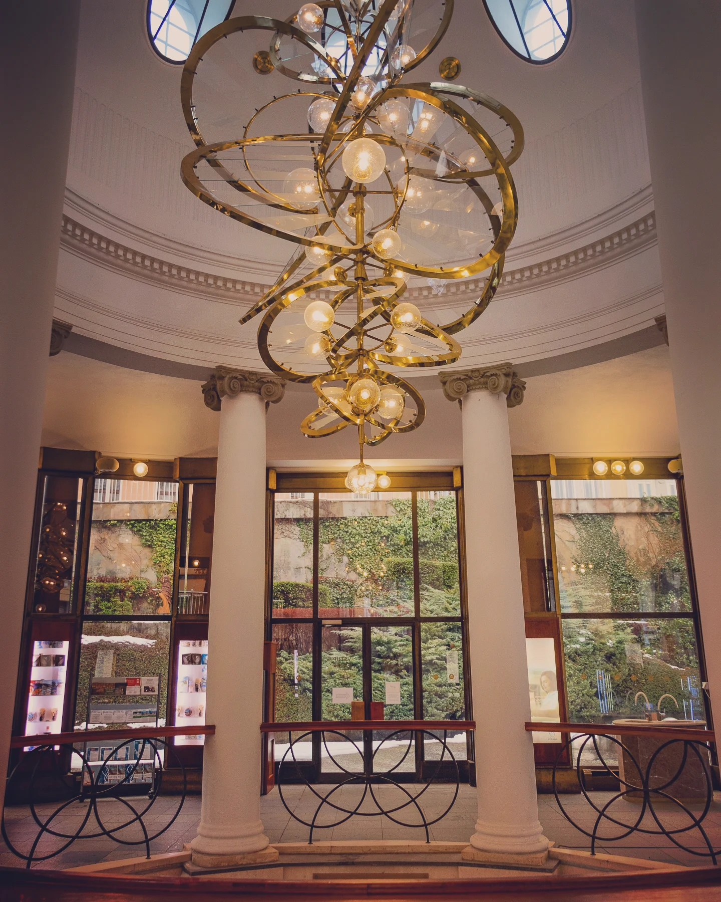 A large modern gold chandelier hanging between white columns inside a rotunda space in the Main Colonnade.