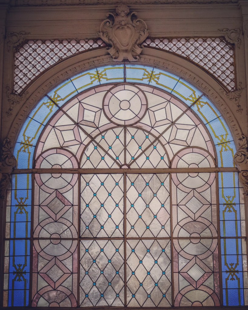 A decorative stained-glass window with geometric patterns in blue, pink, and cream framed by carved stone inside the Main Colonnade in Mariánské Lázně.