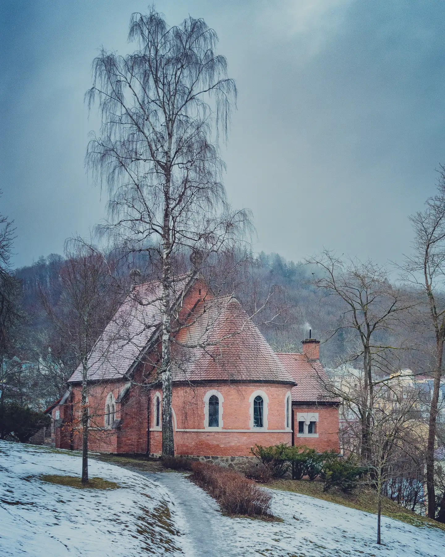 A small red brick chapel on a snowy hillside surrounded by bare trees in Mariánské Lázně.