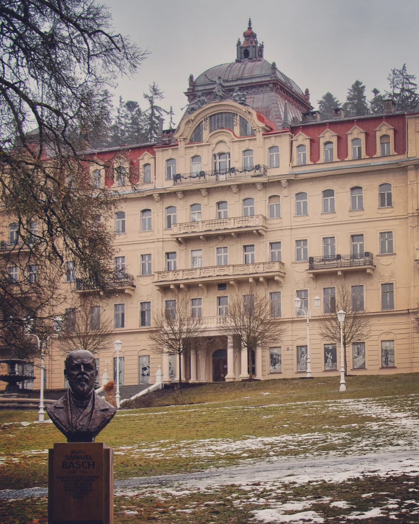 A bronze bust of Samuel Basch standing in front of a large historic spa building in Mariánské Lázně.