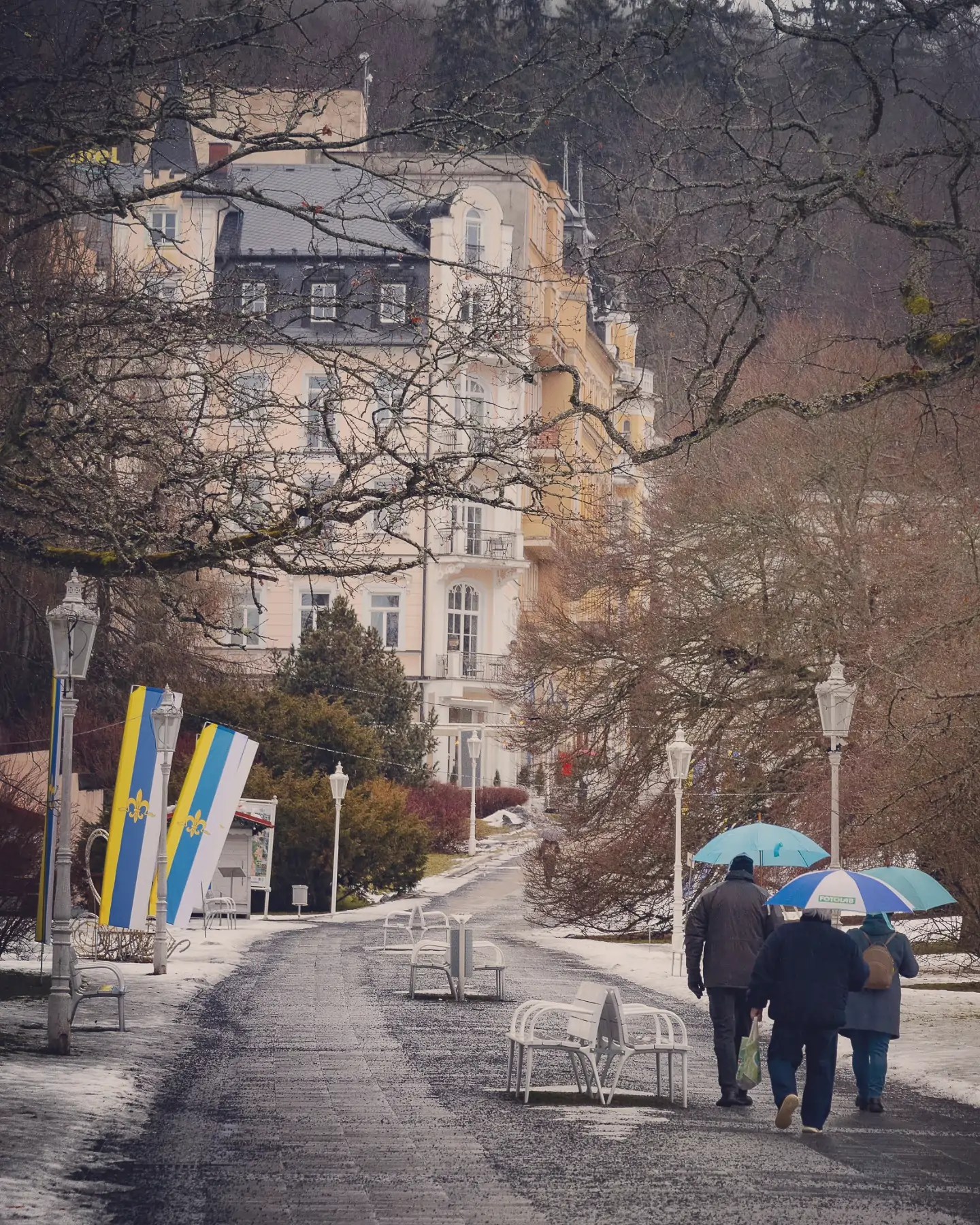 Three people walking with umbrellas along a snowy park promenade lined with historic buildings in Mariánské Lázně.