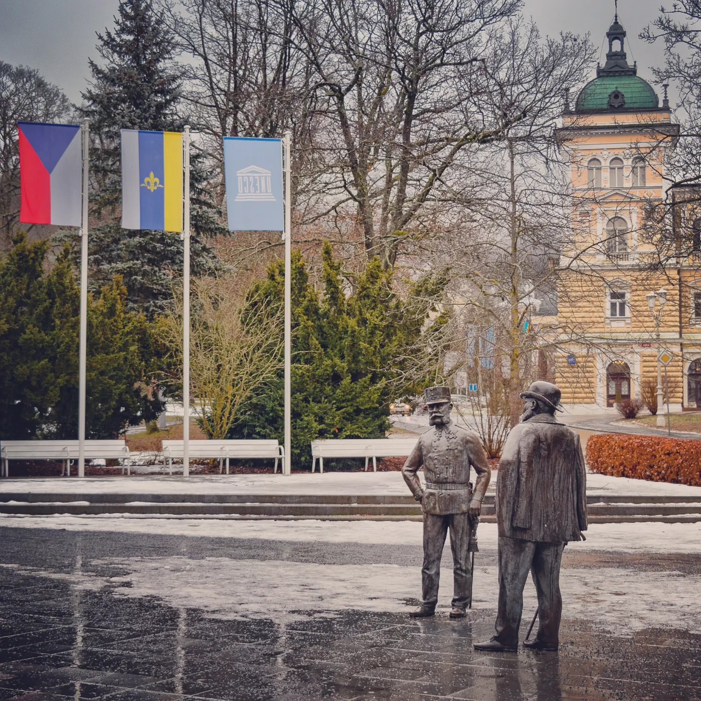 Two bronze statues standing in a square with Czech, local, and UNESCO flags behind them in Mariánské Lázně.