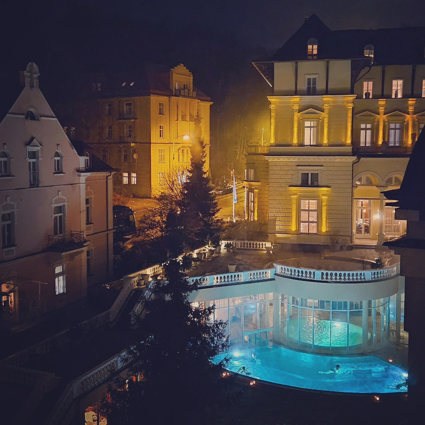 An illuminated outdoor spa pool with turquoise water beneath a historic hotel building lit in golden light at night in Mariánské Lázně.
