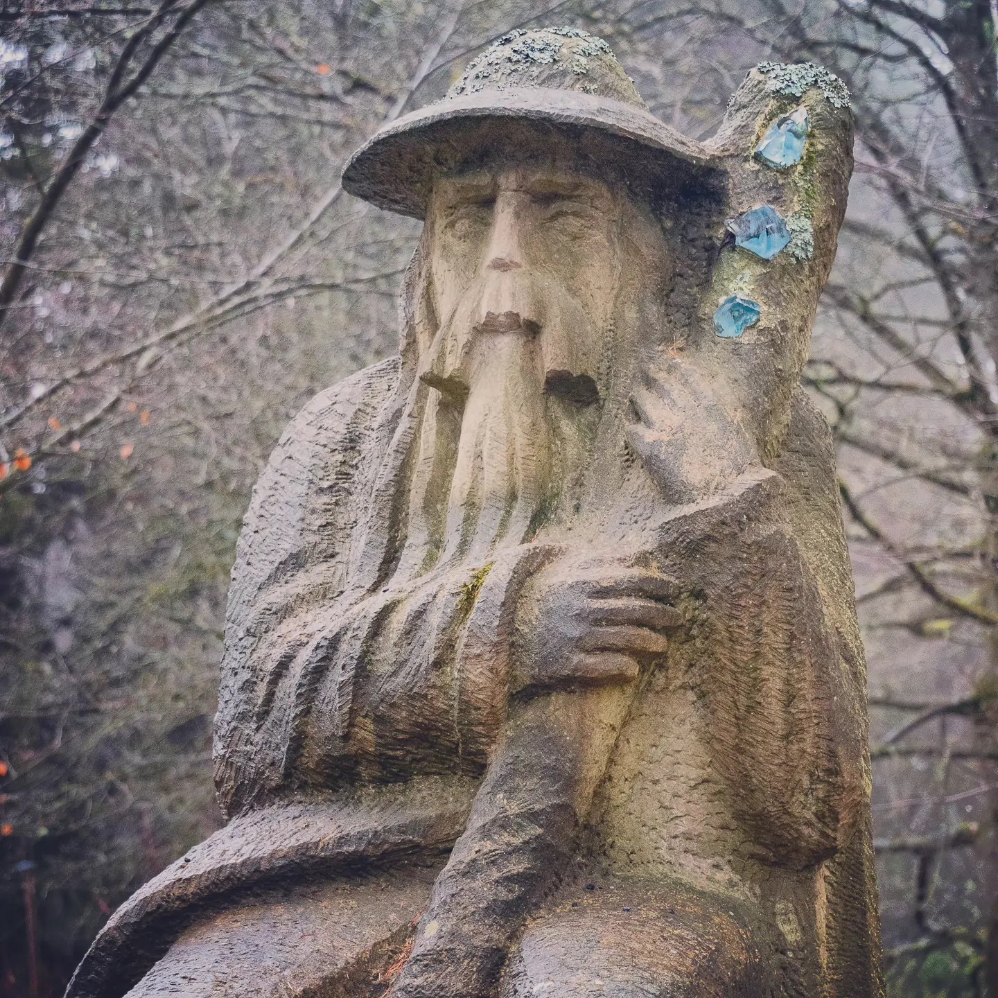 A weathered stone sculpture of a bearded man wearing a wide-brimmed hat and holding a staff in a forested park setting.