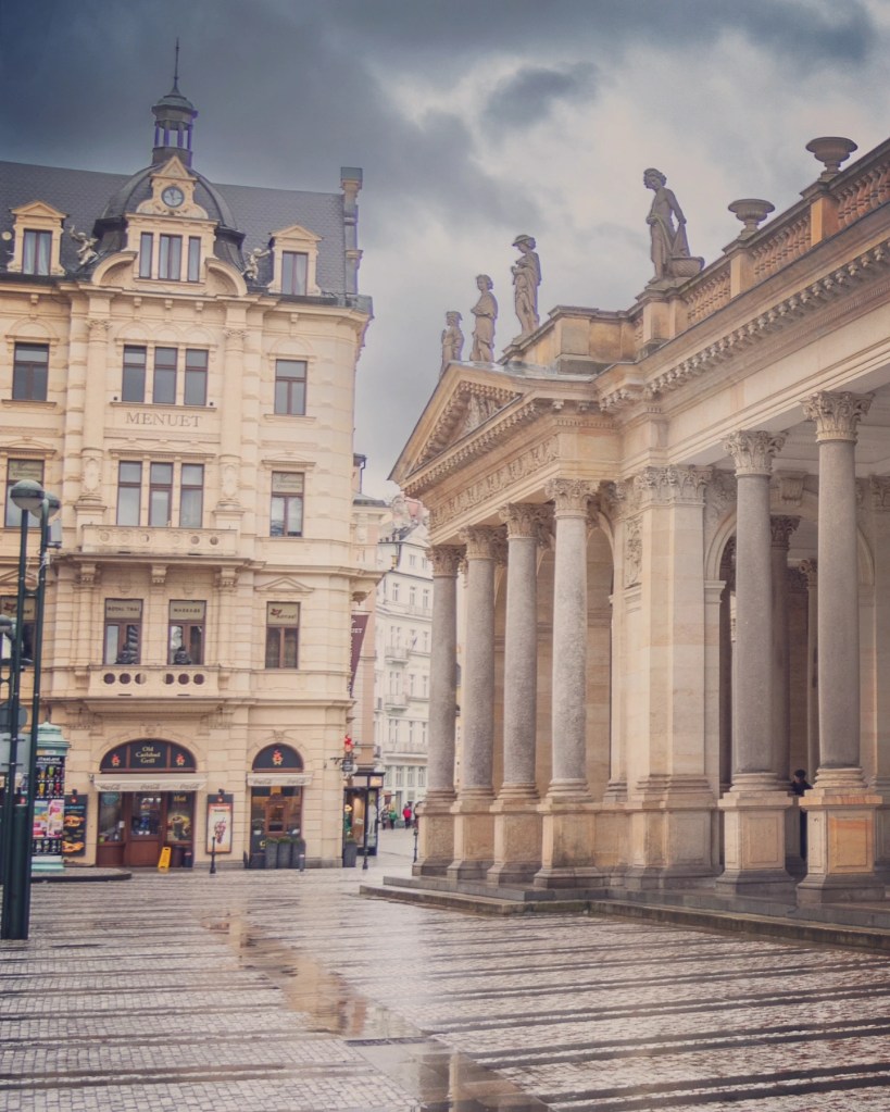 A row of statues stands atop the roofline of a grand colonnade supported by tall Corinthian columns on a wet cobblestone square.