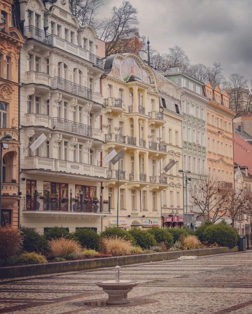 A small stone spring fountain stands in the foreground with ornate pastel spa buildings lining the street behind it.
