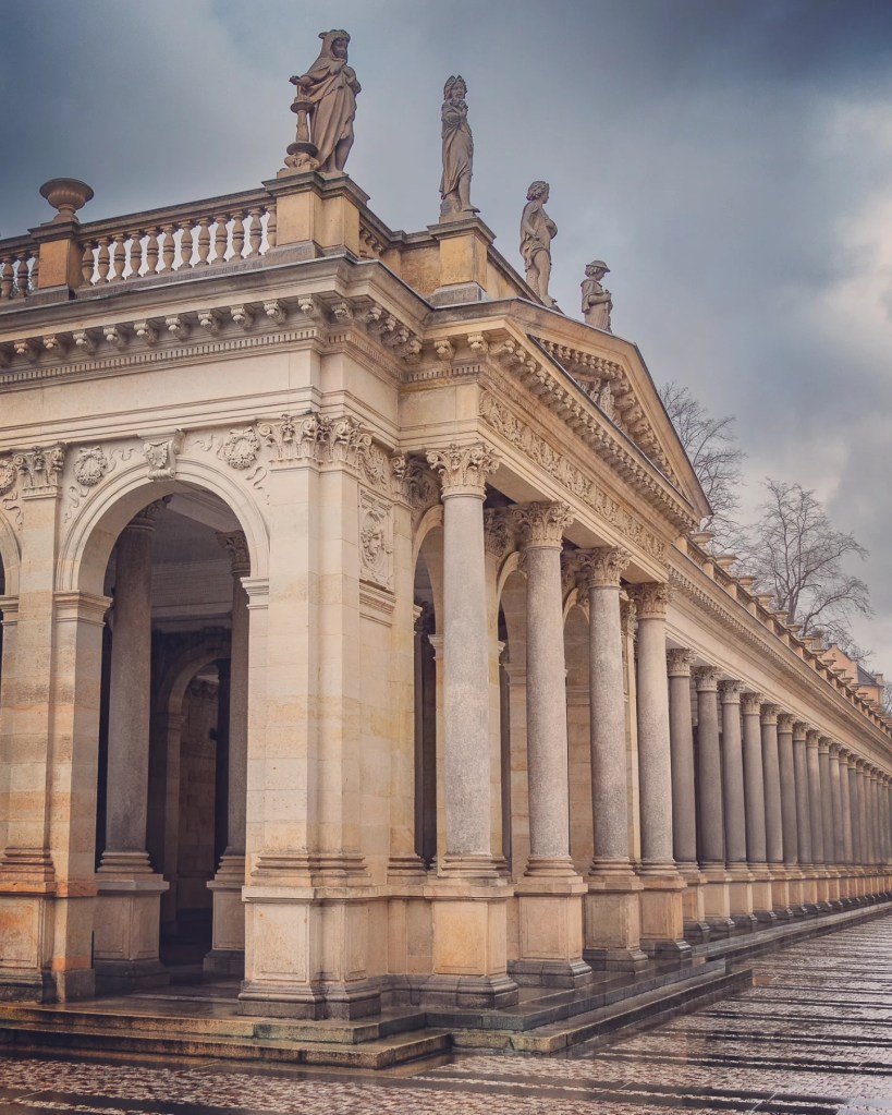 A long row of stone columns with Corinthian capitals stretches into the distance beneath statues along the roof of the Mill Colonnade.