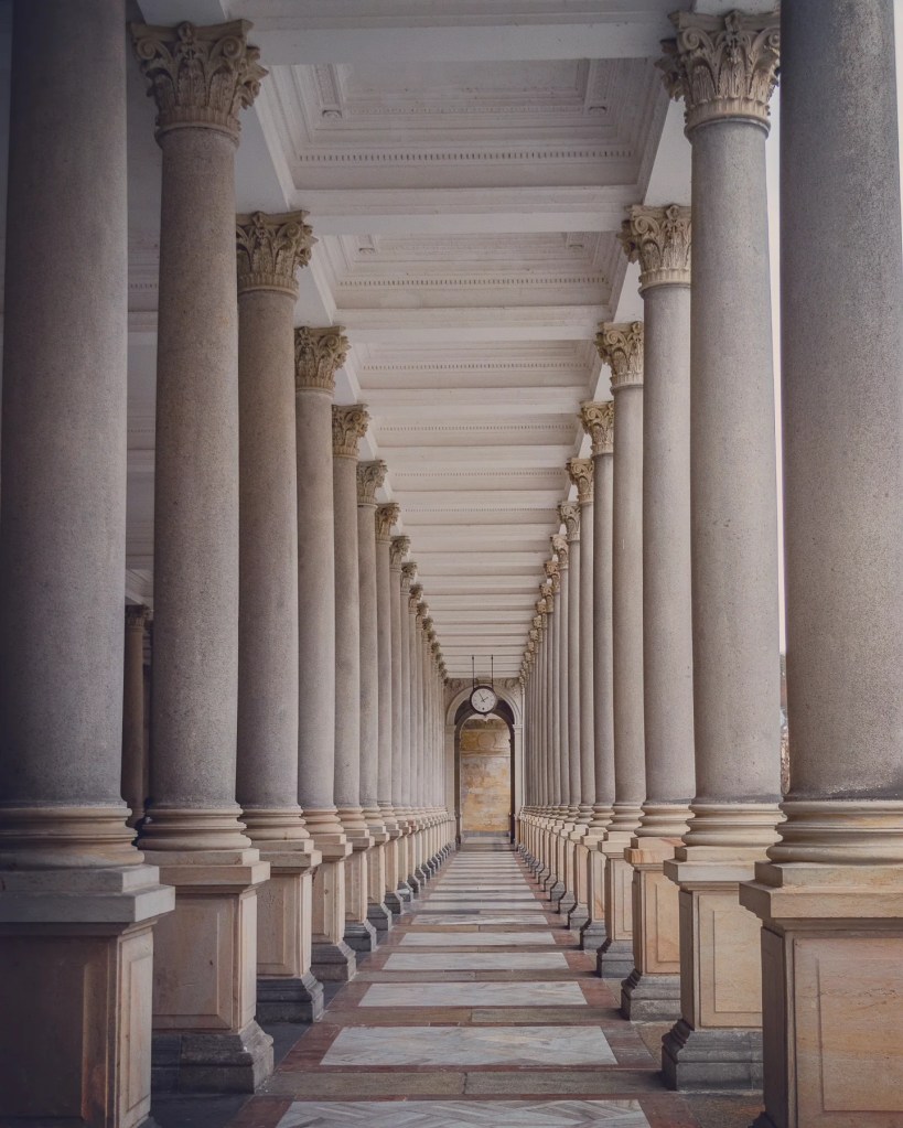 A symmetrical view down the interior corridor of the Mill Colonnade shows rows of tall stone columns leading toward a clock at the far end.