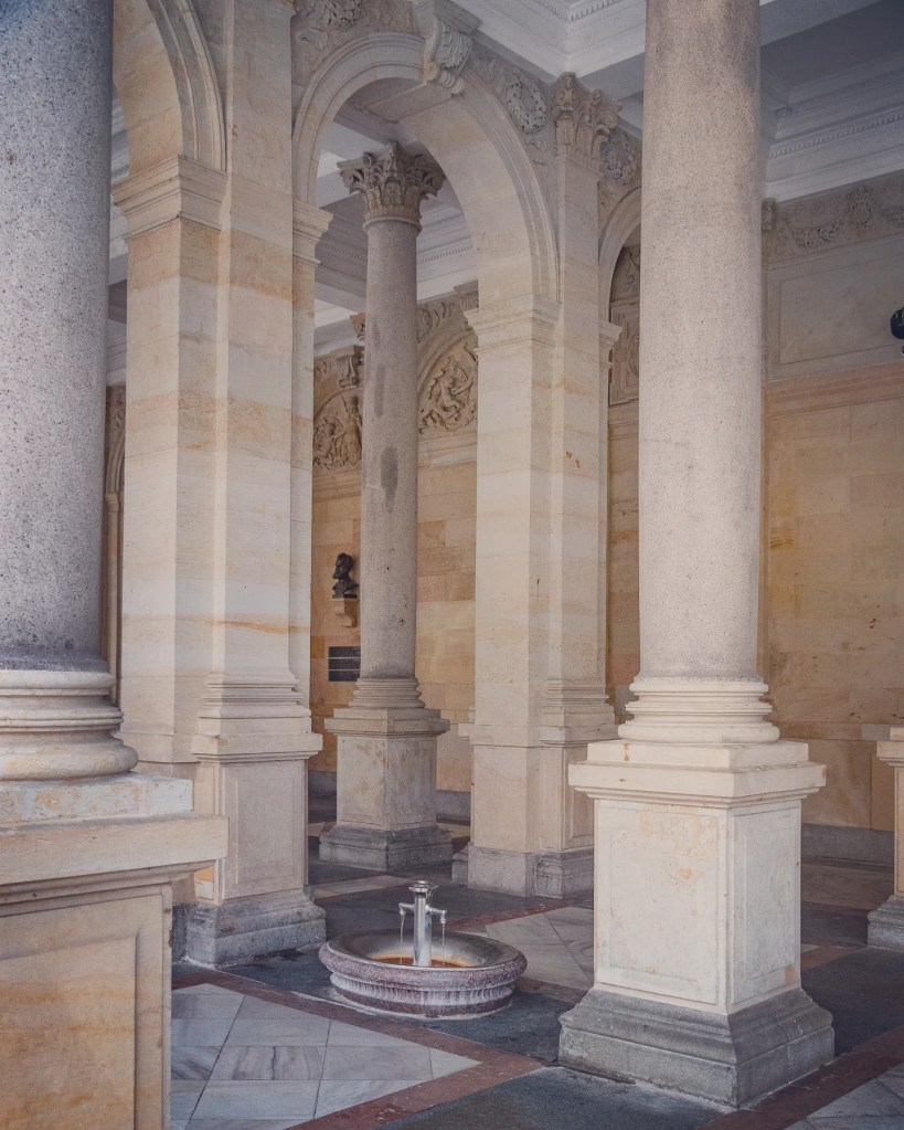 A small mineral spring fountain set in a shallow stone basin beneath tall columns inside the Mill Colonnade in Karlovy Vary.