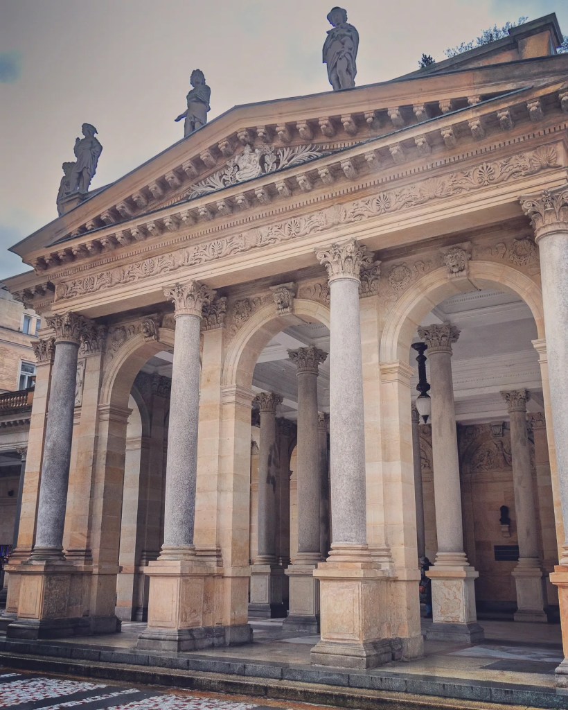 The exterior corner of the Mill Colonnade in Karlovy Vary with tall stone columns, ornate arches, and statues standing along the roofline.