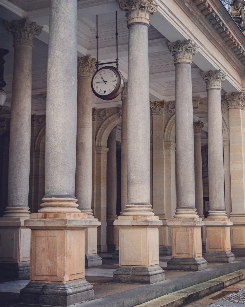 A hanging round clock suspended between rows of tall stone columns inside the Mill Colonnade in Karlovy Vary.