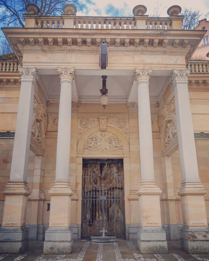 A gated mineral spring set into an ornate stone wall beneath columns at the Mill Colonnade in Karlovy Vary.
