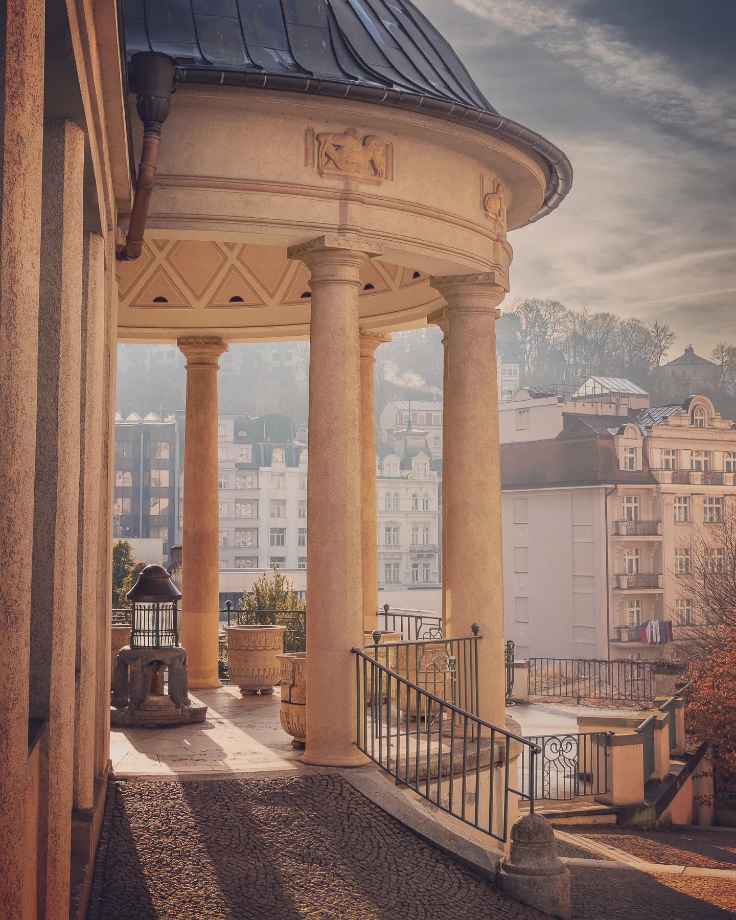 A colonnaded terrace with cream-colored columns overlooking Karlovy Vary’s rooftops in soft morning light.