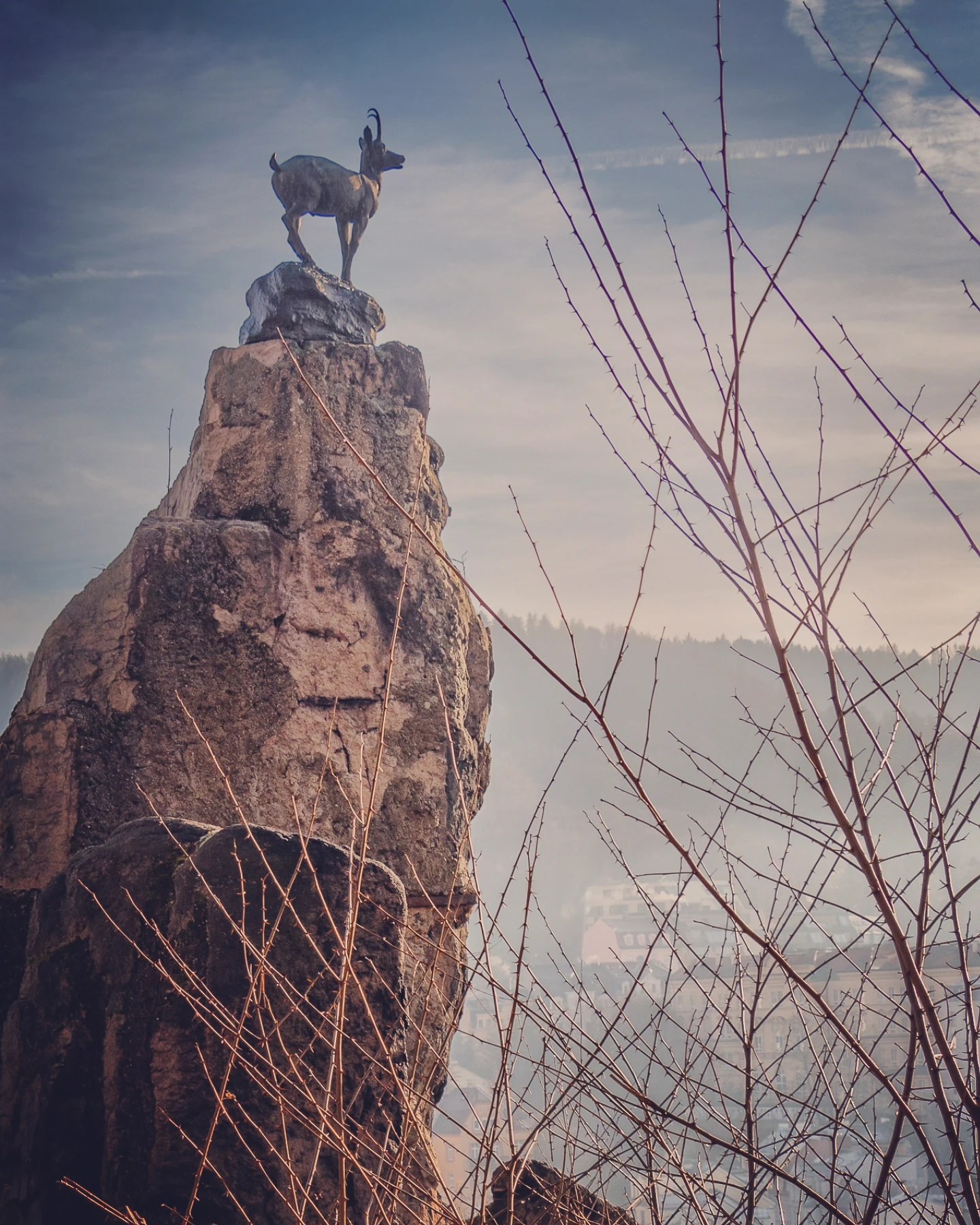 A bronze mountain goat statue standing on top of a tall rock formation overlooking Karlovy Vary.