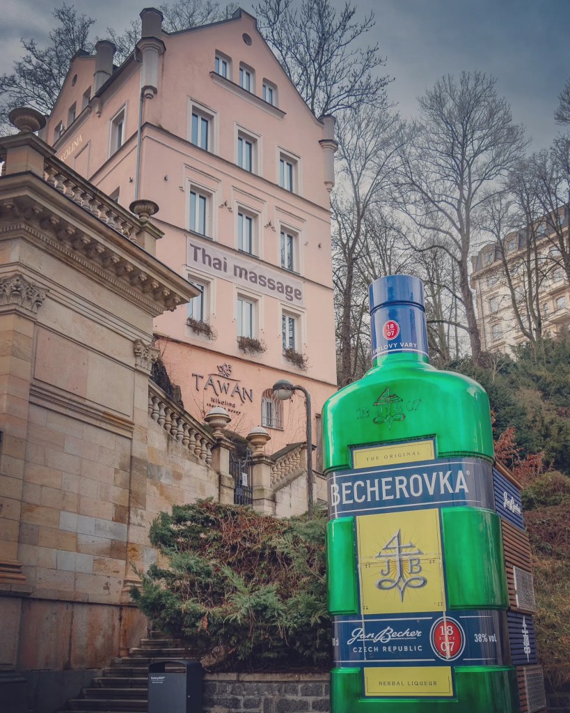 A large green Becherovka bottle sculpture stands beside stone steps and historic buildings in Karlovy Vary.