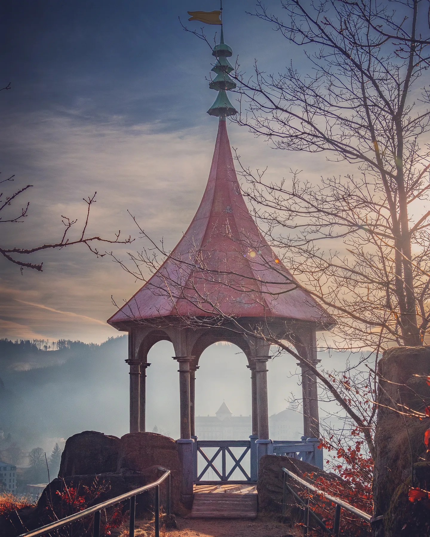 A small red-roofed pavilion stands on a hill overlooking Karlovy Vary in soft morning light.