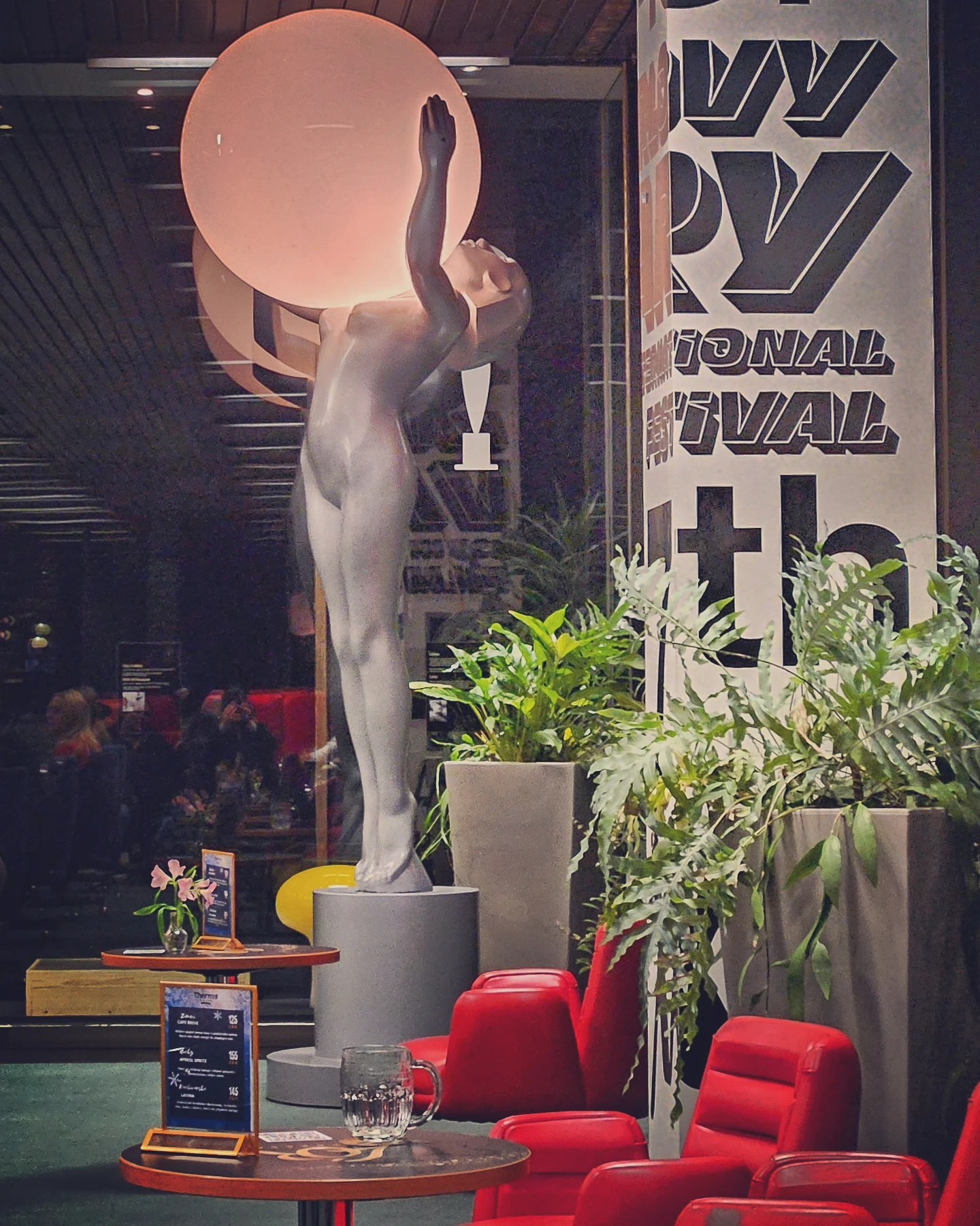 A modern statue of a nude female figure holding a glowing sphere stands inside a building with red chairs nearby.