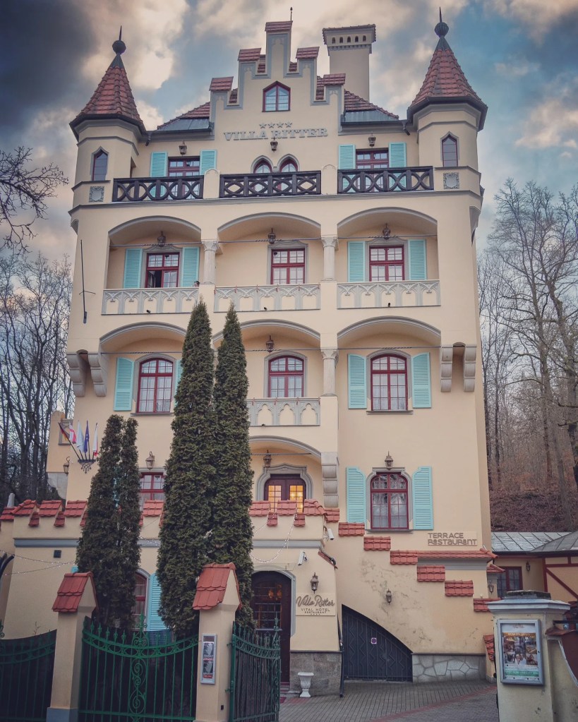 A pale yellow historic building labeled “Villa Ritter” with turrets and balconies rises behind tall evergreen trees.