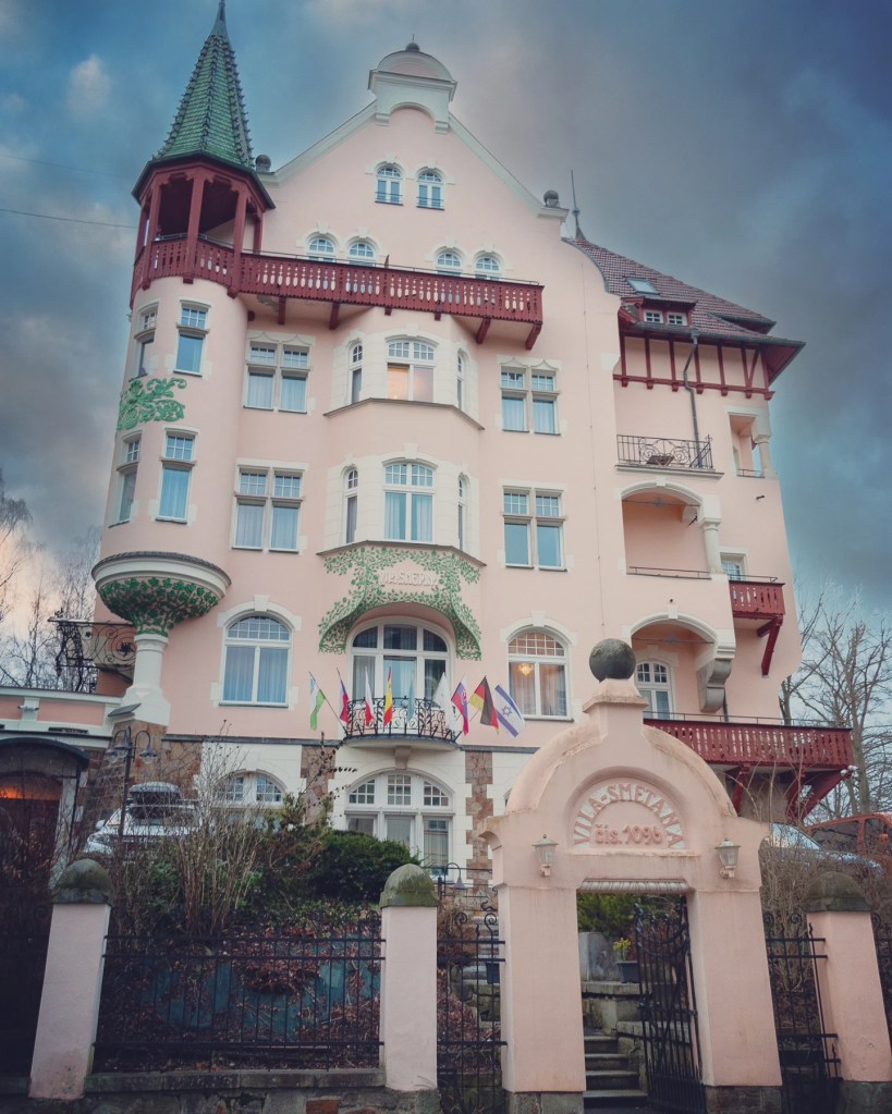 A pale pink historic villa with a turreted corner tower, red wooden balconies, and flags hanging above the entrance in Karlovy Vary.