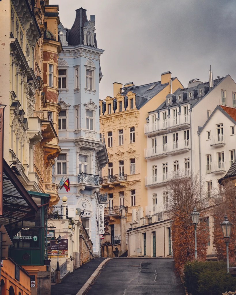 A narrow uphill street lined with pastel-colored historic buildings featuring ornate façades and balconies in Karlovy Vary.