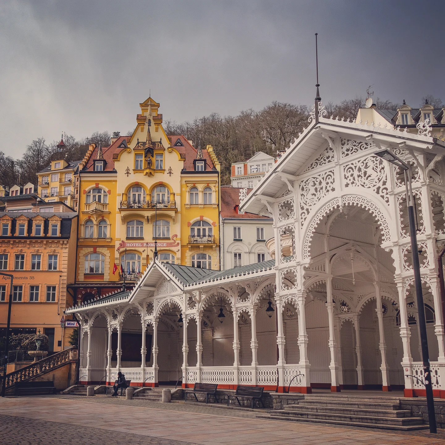The white wooden Market Colonnade in Karlovy Vary stands in front of pastel spa buildings and a wooded hillside.