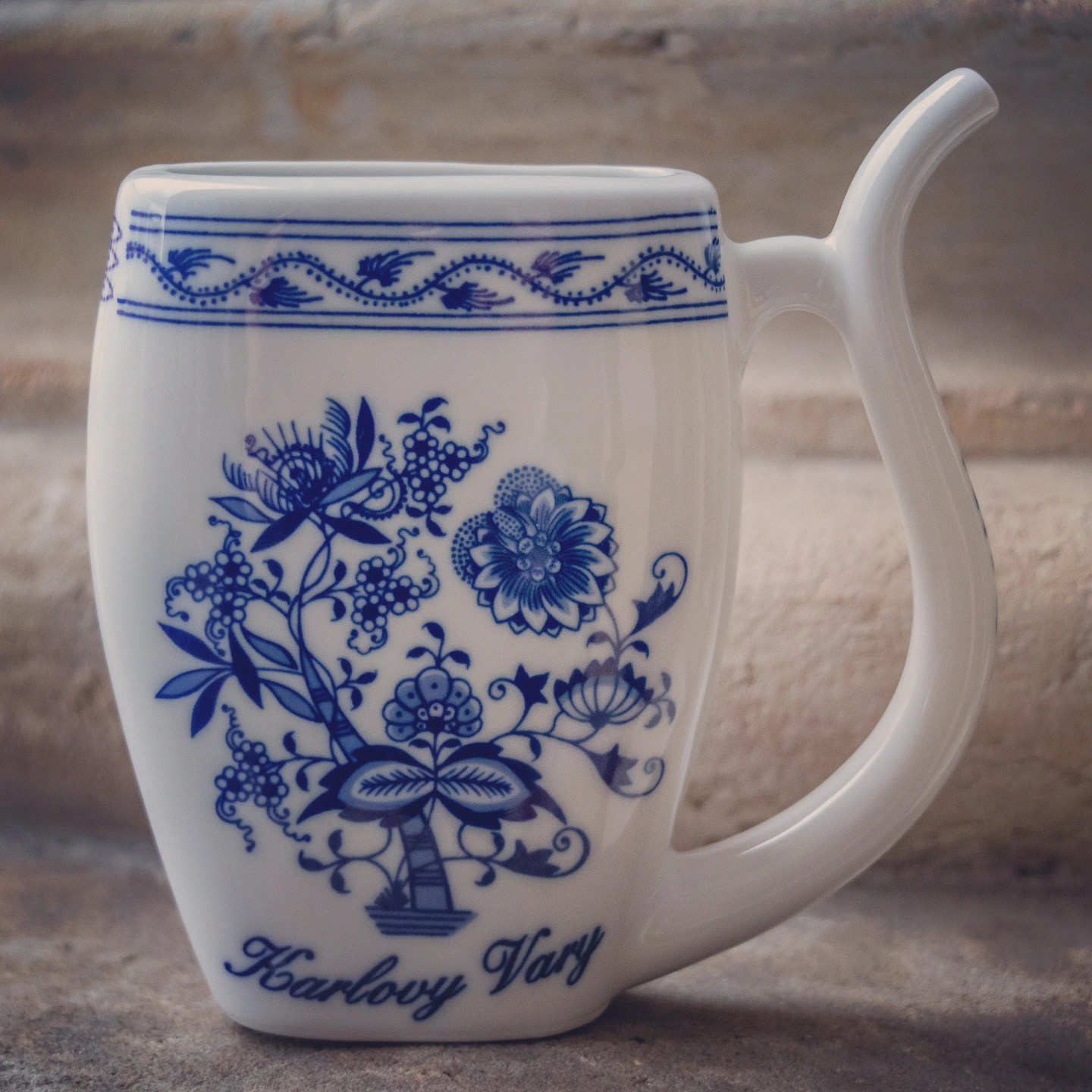 A traditional white porcelain spa cup with a curved drinking spout and blue floral pattern labeled Karlovy Vary.