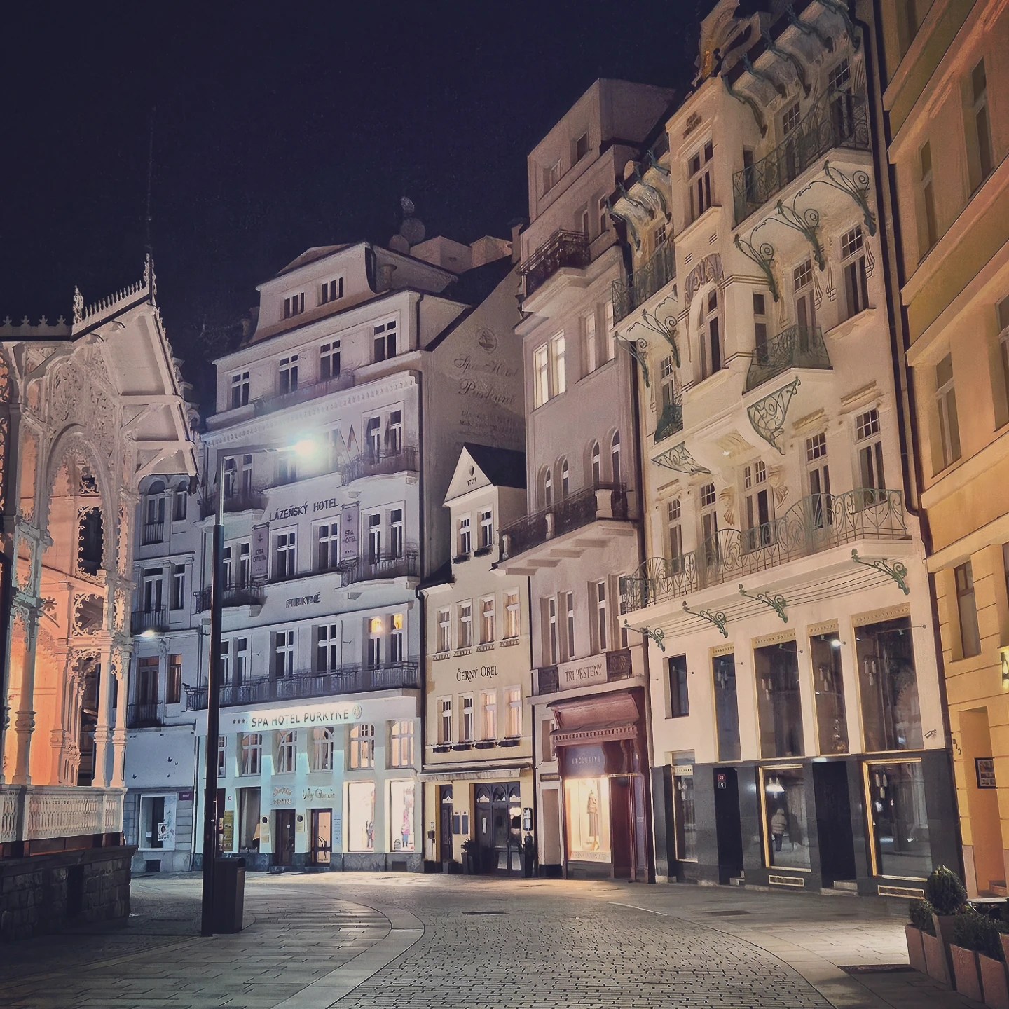 Historic buildings lit at night beside the Market Colonnade in Karlovy Vary along a curved cobblestone street.