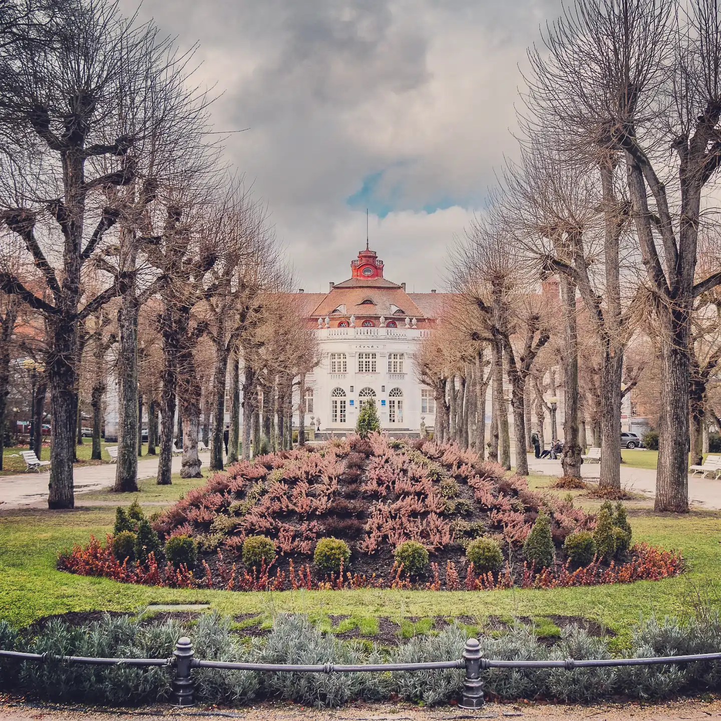 A formal park garden with trimmed trees lining a central flower bed and a white historic building at the end in Karlovy Vary.