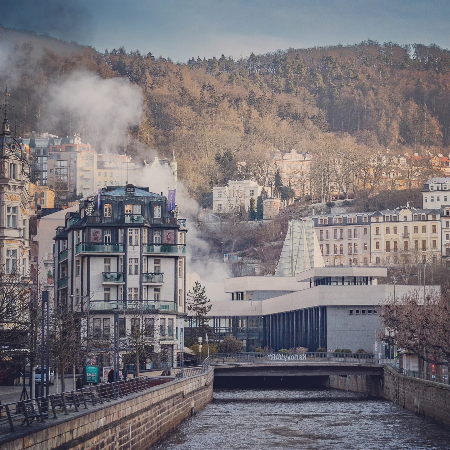Steam rising along the Teplá River in Karlovy Vary with historic buildings and wooded hills behind.
