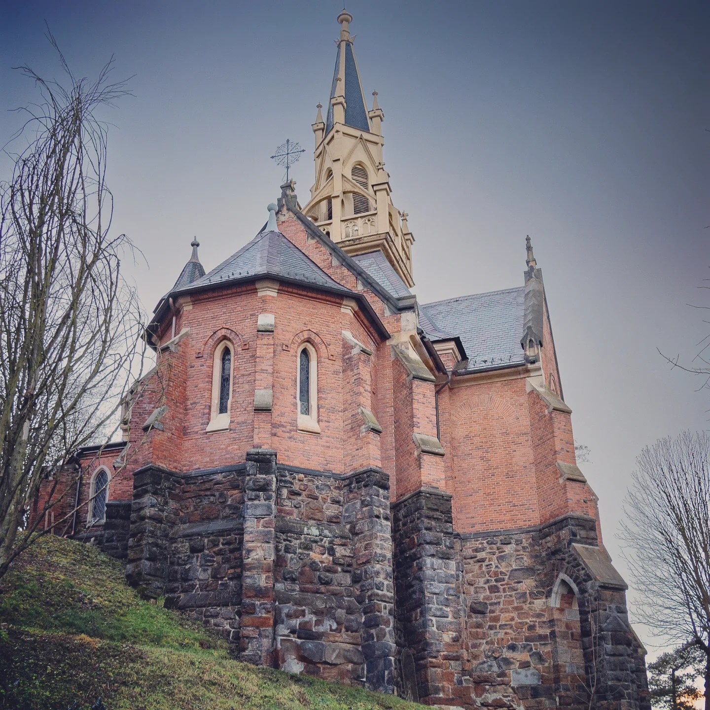 A red brick Gothic-style church with a tall pointed spire stands on a stone foundation on a grassy hillside in Karlovy Vary.